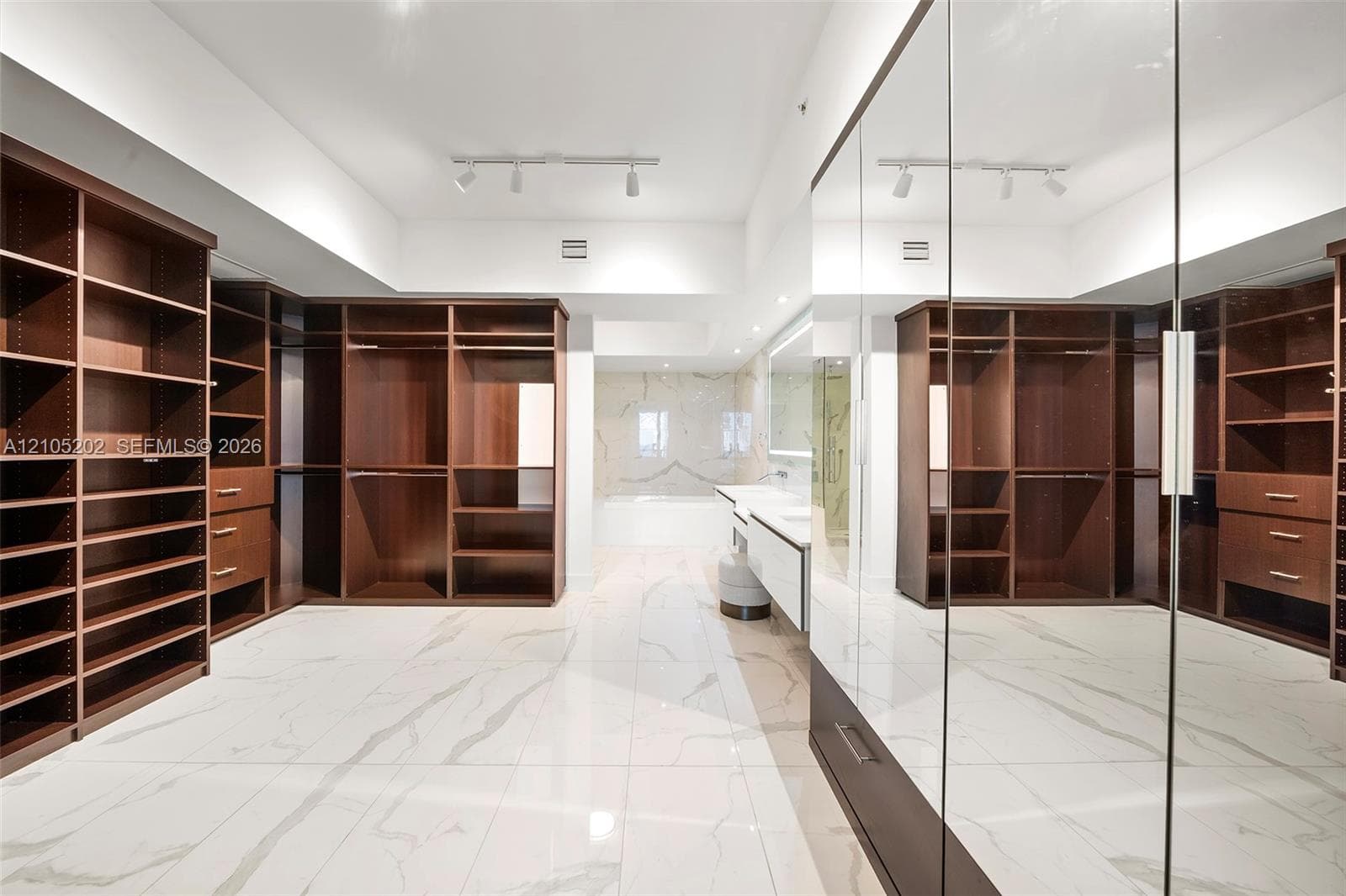 Custom walk-in dressing area featuring extensive dark wood-finish cabinetry, floor-to-ceiling mirrored closet doors, polished stone-finish flooring with vein detailing, and recessed track lighting