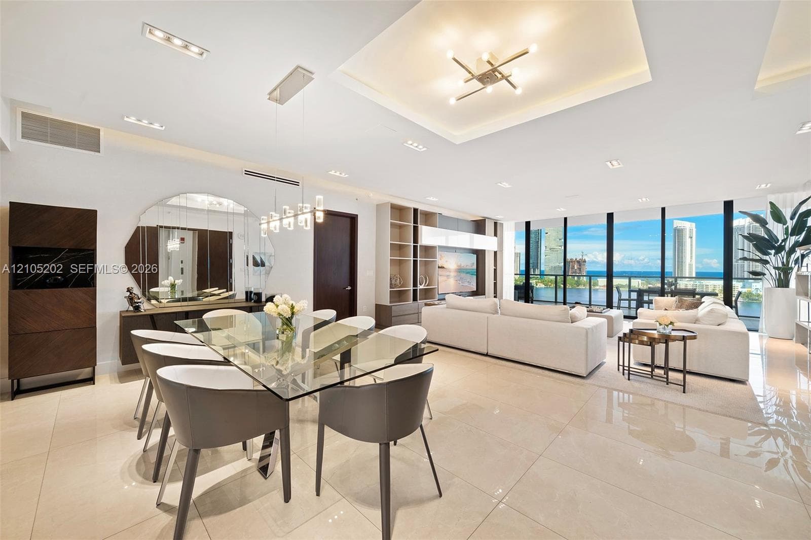 Open-concept interior featuring large-format tile flooring, tray ceilings with recessed lighting, custom built-in cabinetry, and floor-to-ceiling windows with water-centric scenery