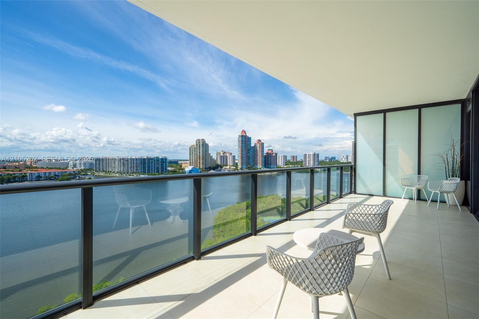 Covered balcony featuring oversized tile flooring, a glass panel railing with a dark metal frame, and water frontage