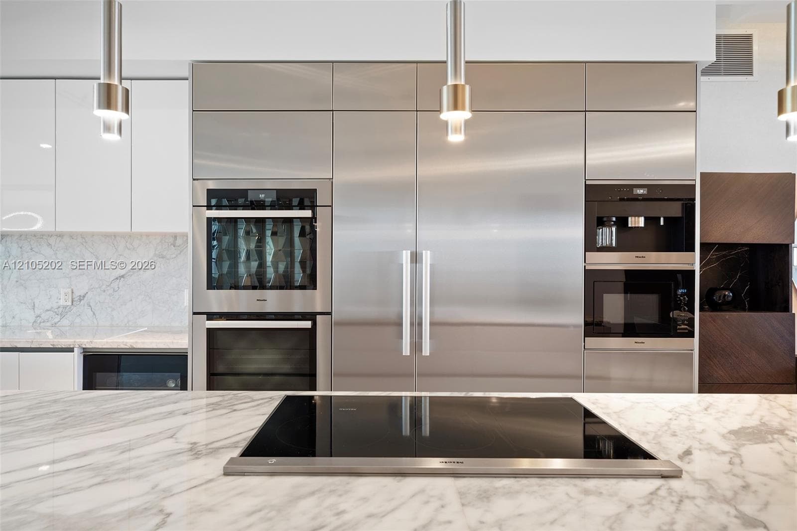 Modern kitchen featuring integrated stainless steel appliances, stone slab countertops, a built-in induction cooktop, high-gloss cabinetry, and pendant lighting