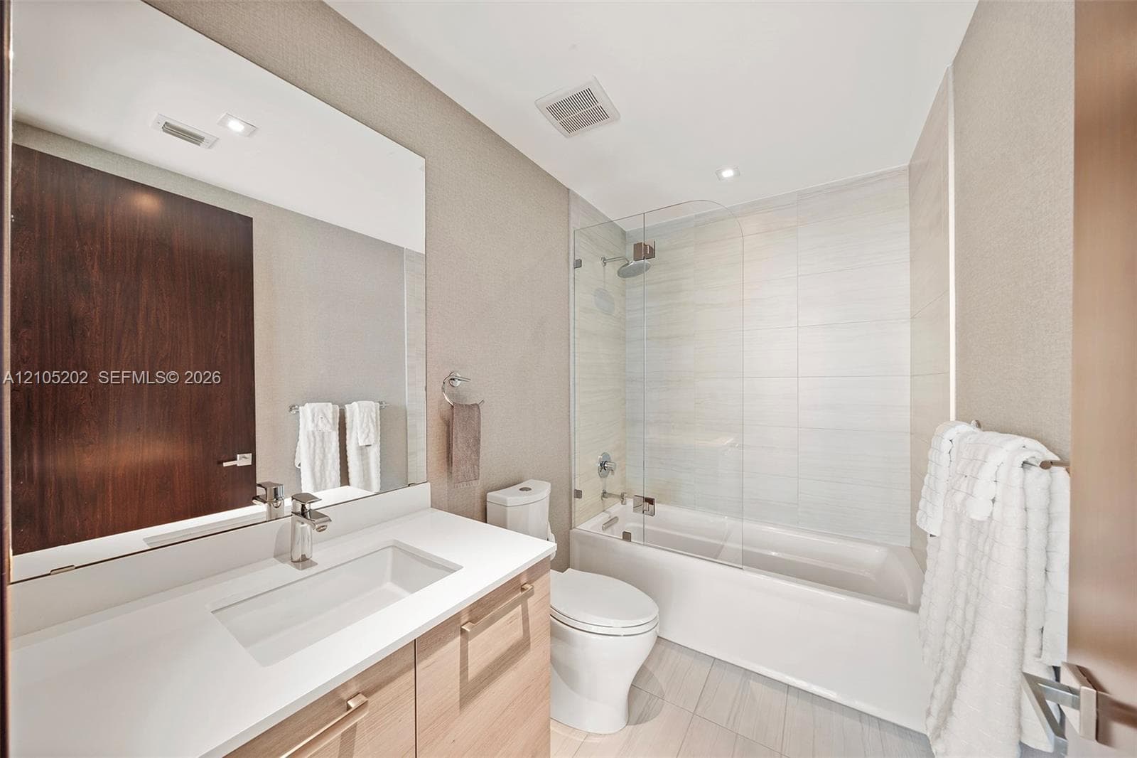 Bathroom featuring a vanity with a white countertop and rectangular undermount sink, large wall-mounted mirror, light-toned wood-finish cabinetry, and a shower-tub combination enclosed by a glass panel with horizontal tile surround