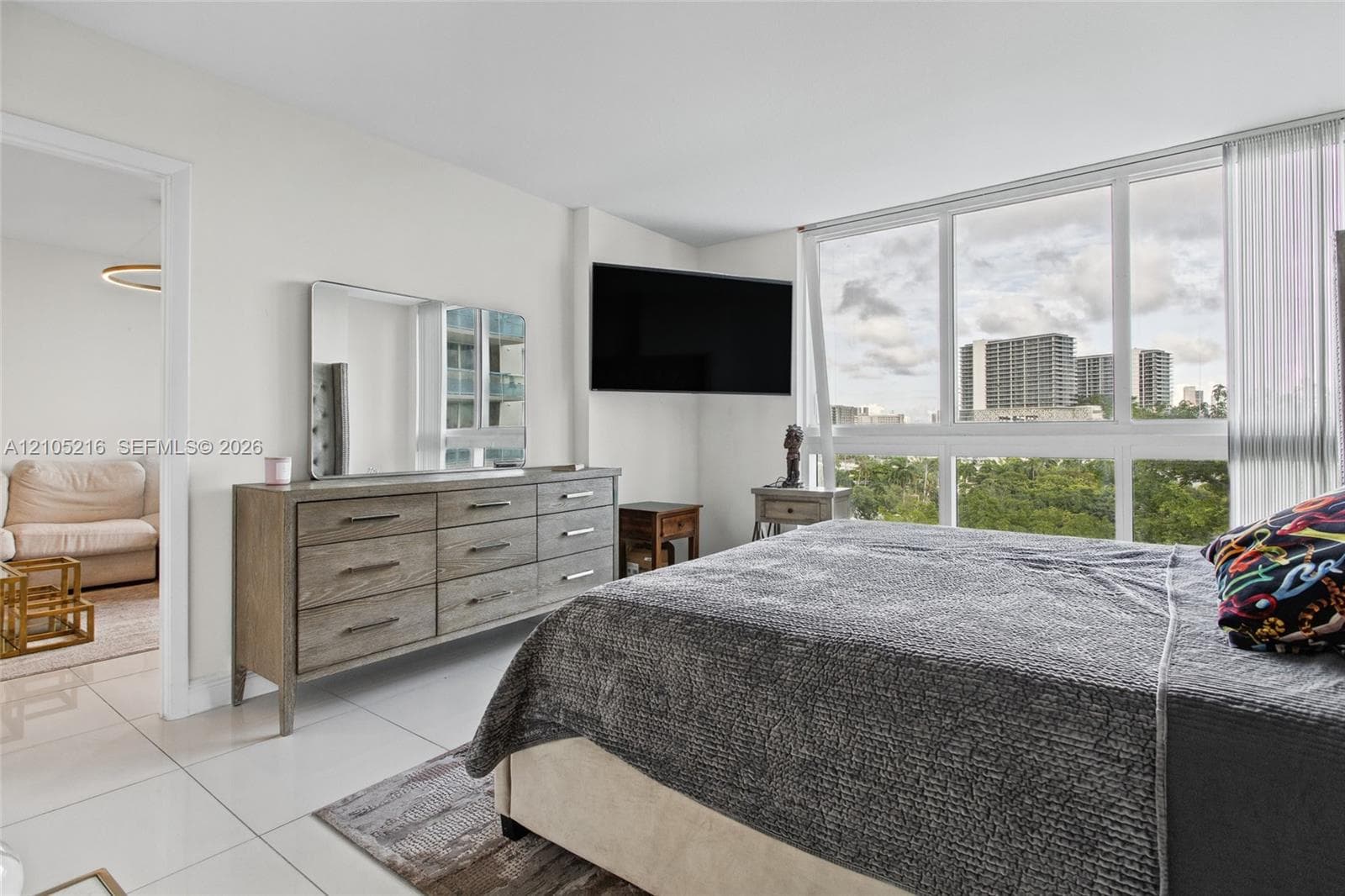 Bright corner bedroom with expansive windows framing cityscape panoramas, light-filled flooring, wood-finish dresser with mirror, wall-mounted flat-screen television, and layered window treatments including sheer panels and vertical blinds
