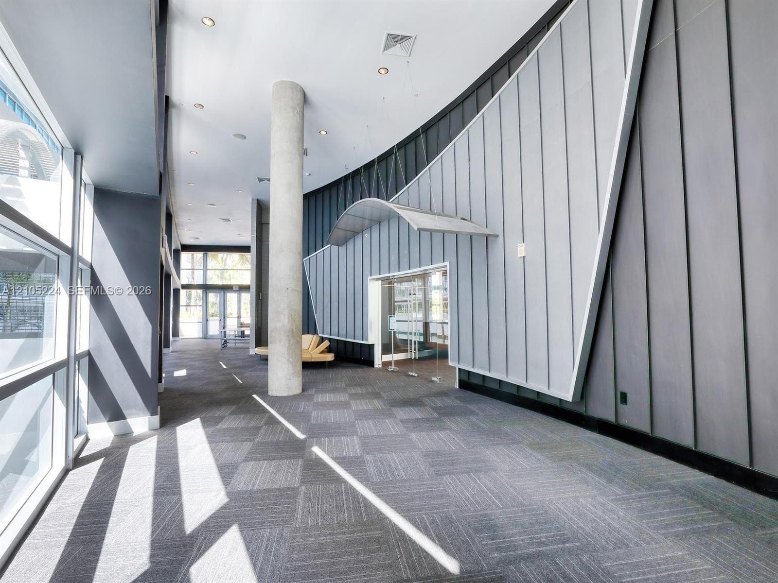 Expansive hallway with full-height windows, patterned carpet flooring, a concrete column, and a distinct curved wall design