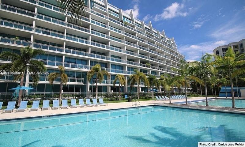Multi-story building with a white facade and glass-front balconies, expansive swimming pool, surrounding paved deck, and mature palm trees