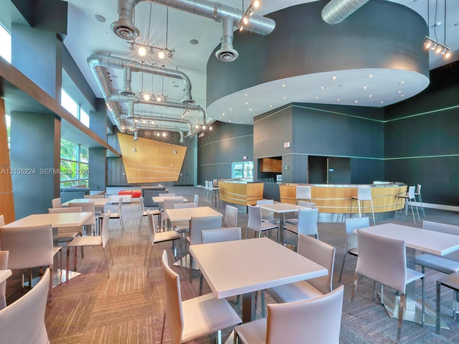 Open communal area featuring exposed ductwork, recessed lighting, a built-in bar with wood-finish paneling, floor-to-ceiling windows, and patterned carpet flooring