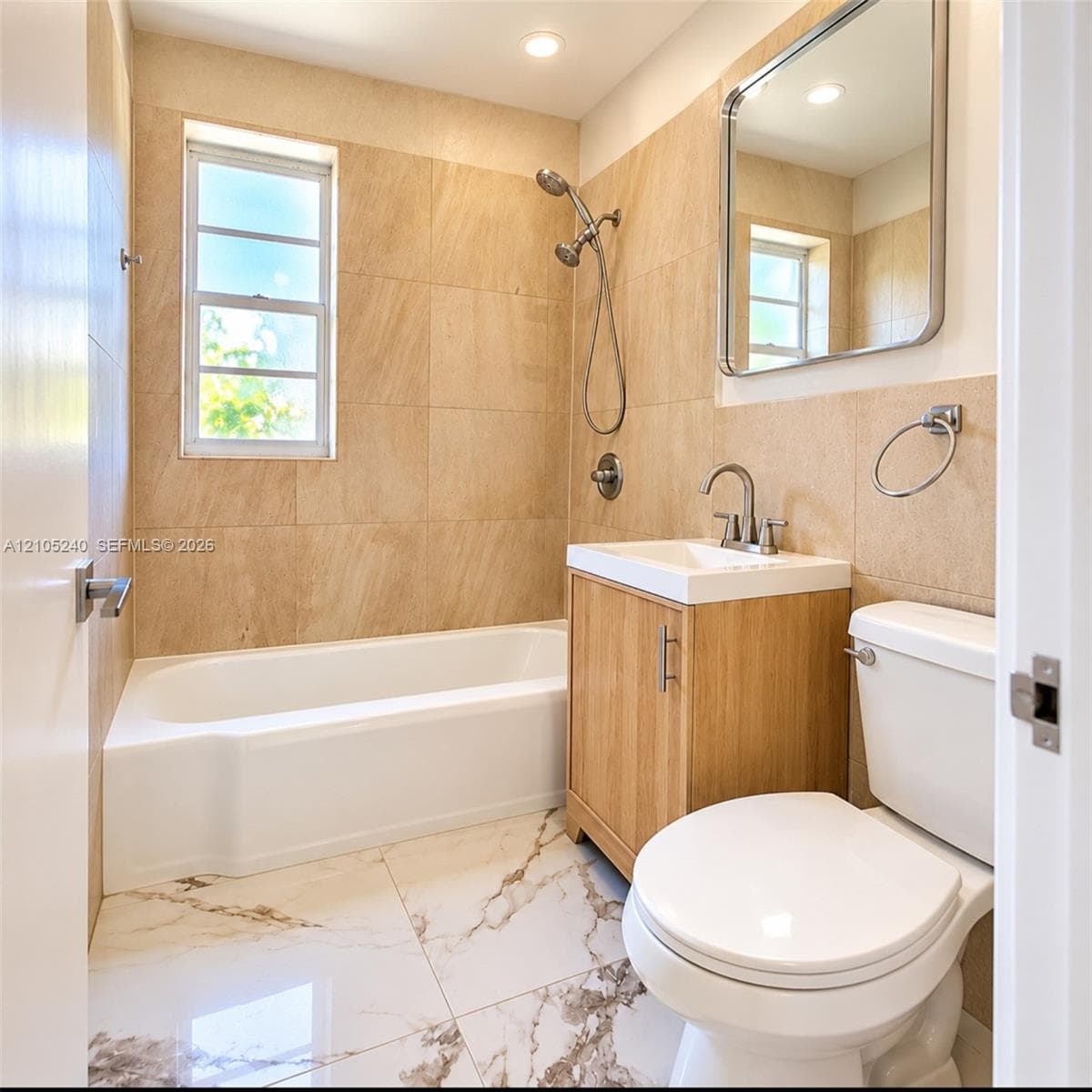 Bathroom featuring large-format wall tiling, a white tub-shower combination, a wood-finish vanity with an integrated basin, and polished tile flooring with veining detail