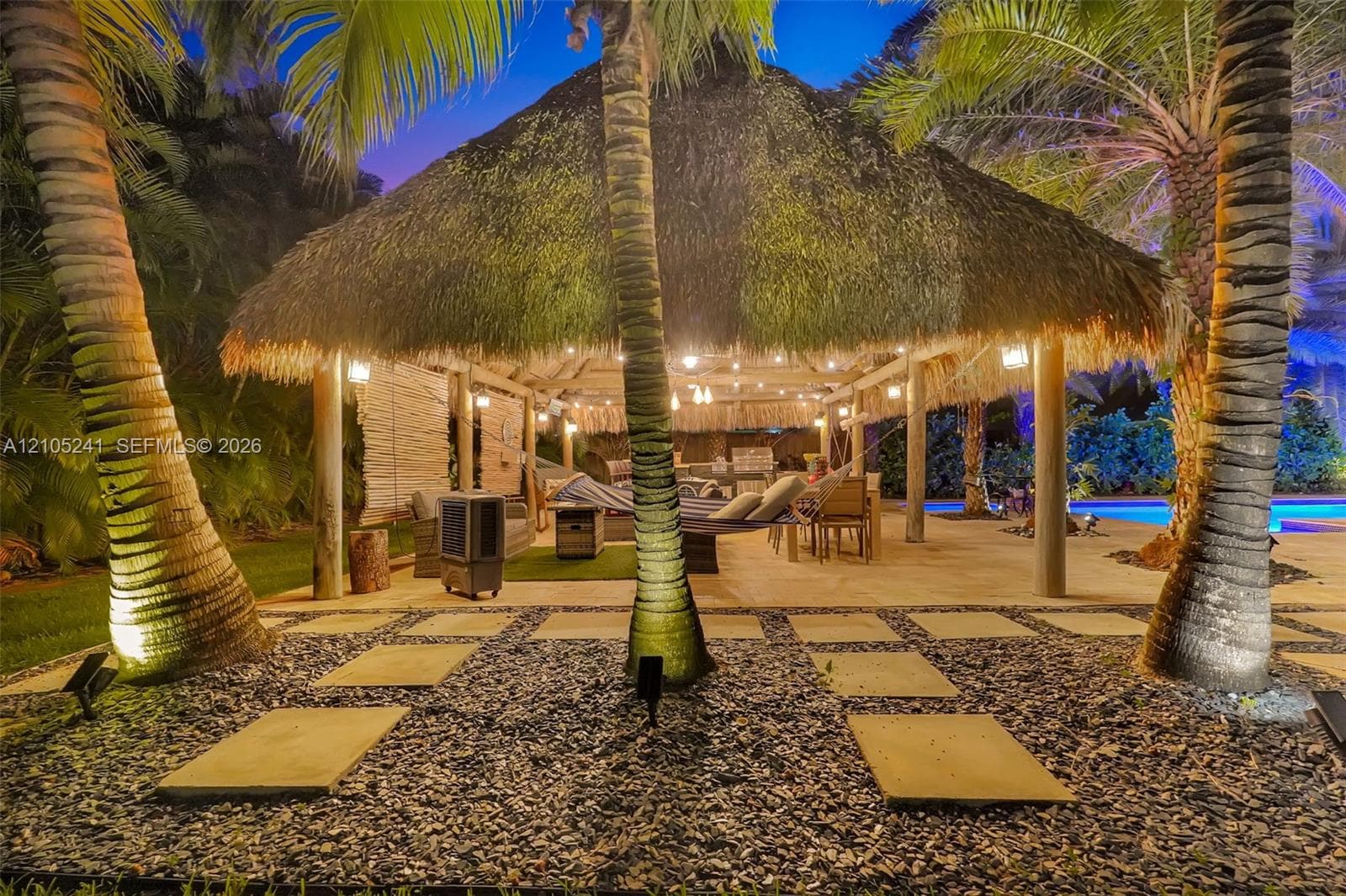 Thatch-roofed outdoor structure with string lighting, surrounded by mature palm trees, featuring a stone paver patio and a gravel walkway with stepping stones