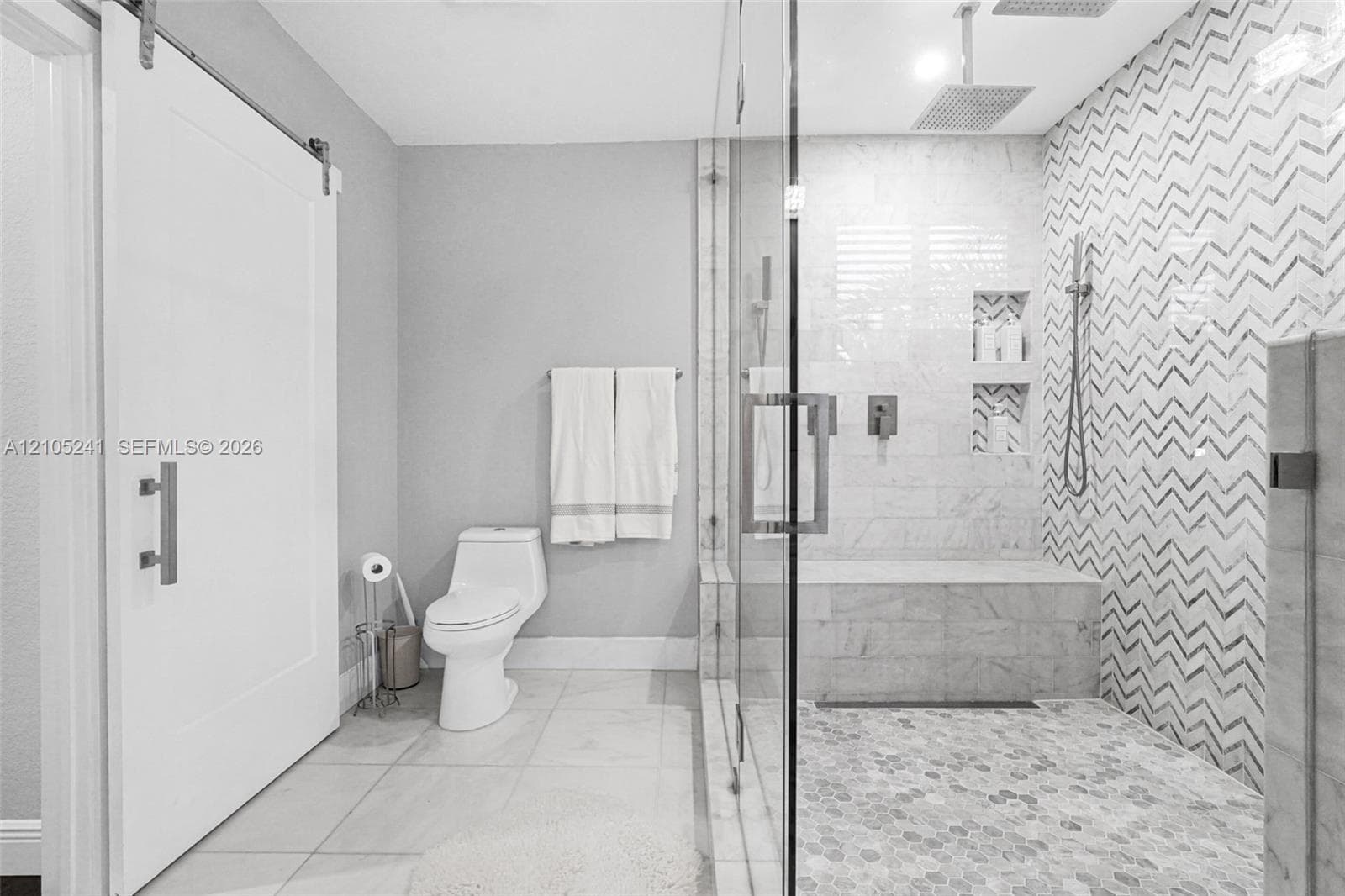 Modern bathroom featuring a sliding barn door, a spacious shower with glass enclosure, mosaic chevron tile accent wall, built-in shower bench, and recessed shower niches