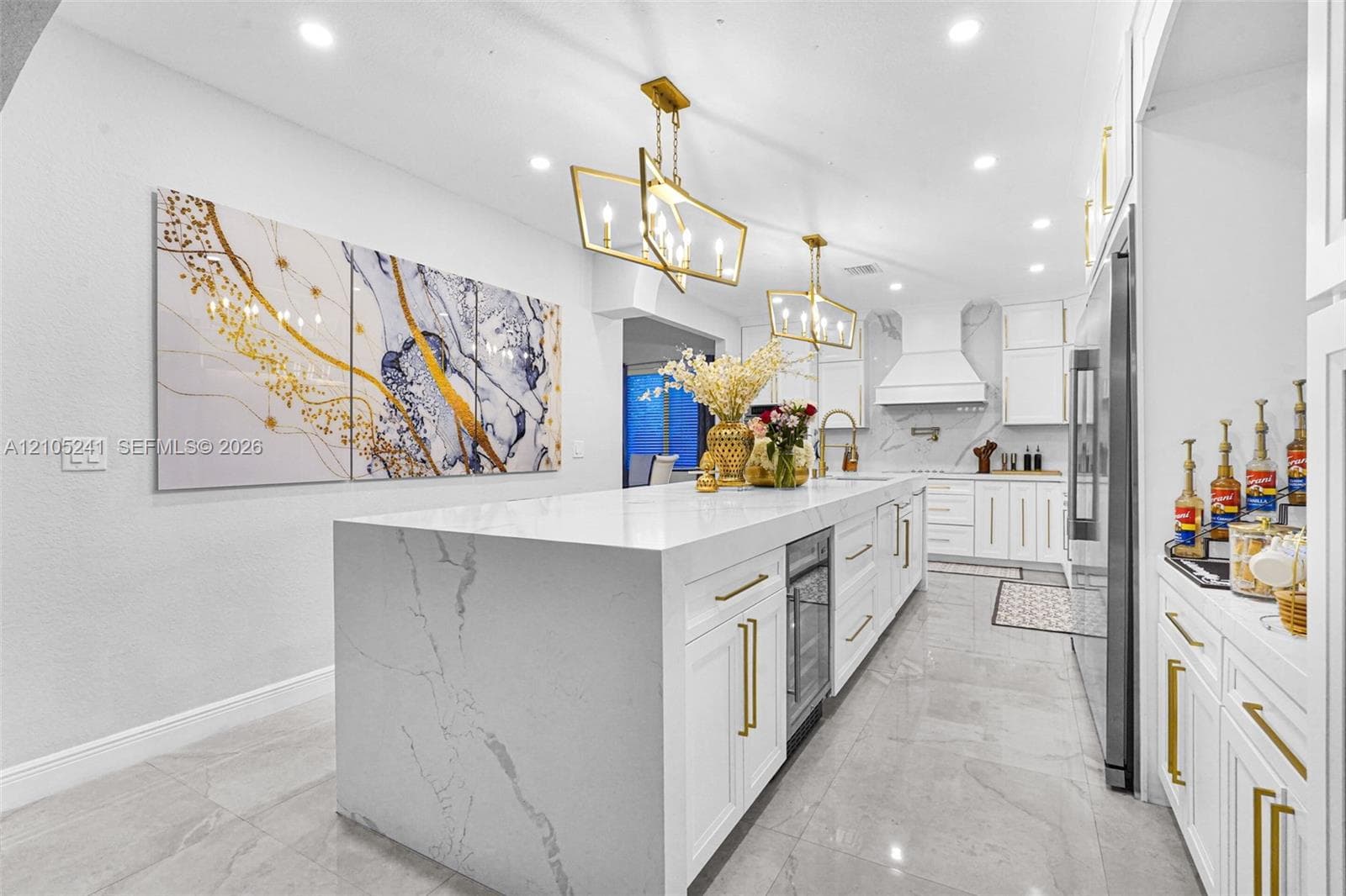 Expansive kitchen featuring an oversized island with a beverage refrigerator, white cabinetry with gold-tone hardware, and recessed lighting