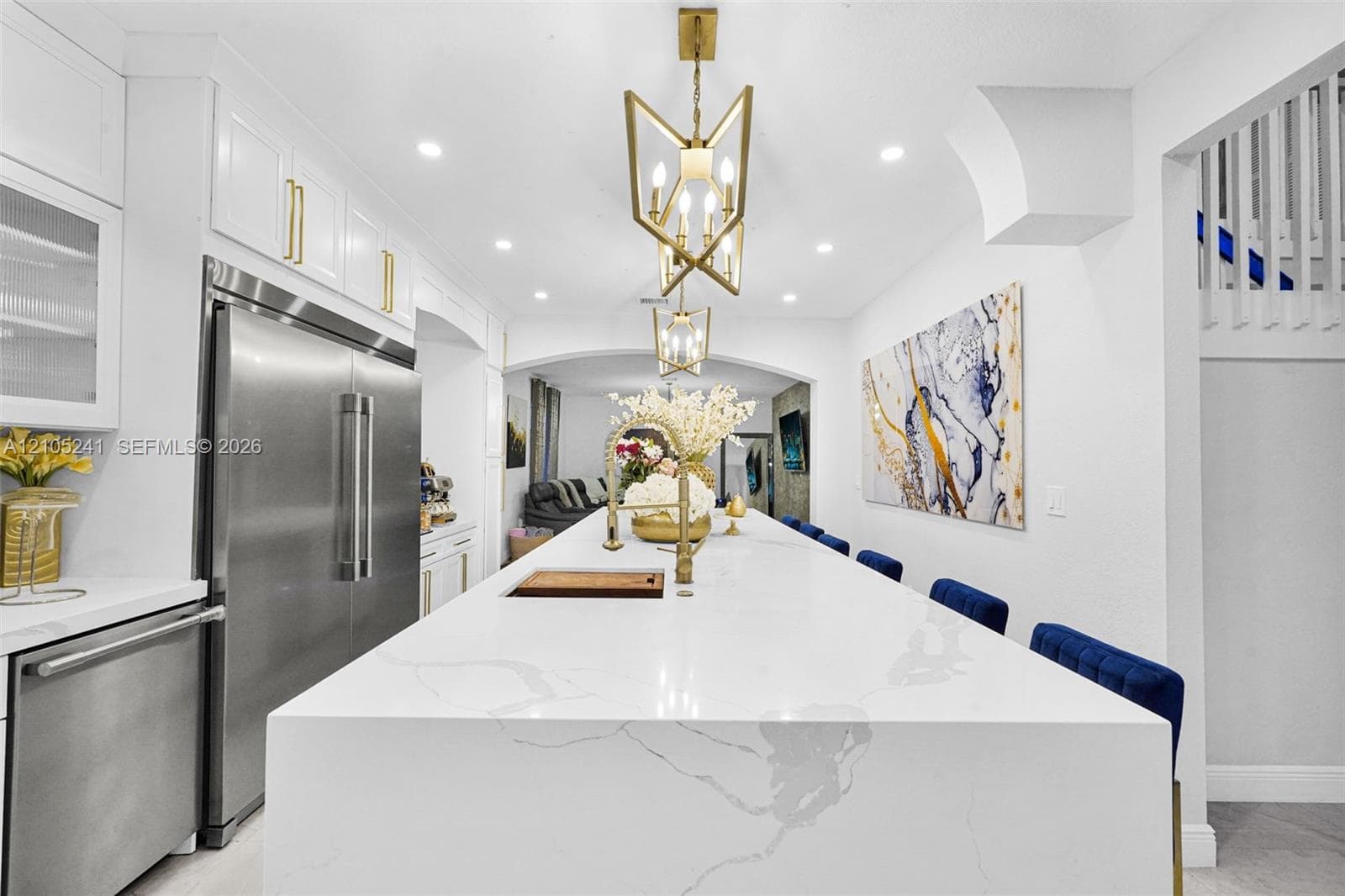 Expansive kitchen island with a waterfall edge, stainless steel refrigerator, white cabinetry with gold-tone hardware, and recessed lighting