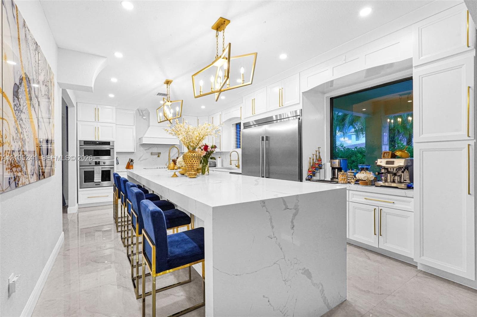 White cabinetry with gold-tone hardware, large center island with waterfall edge, built-in double oven, stainless steel refrigerator, and recessed lighting