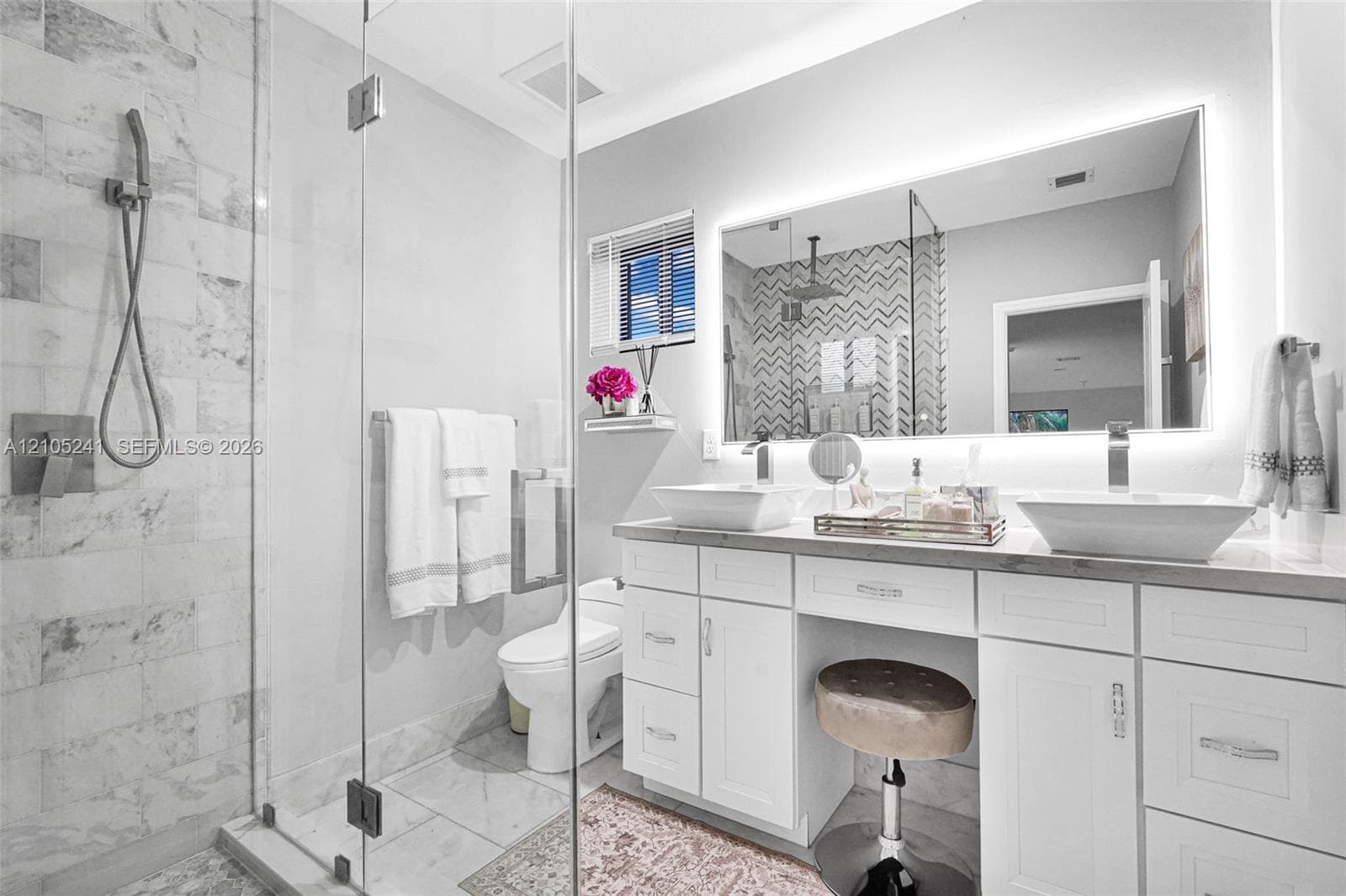 Glass-enclosed shower with tiled walls, dual vessel sinks, vanity with integrated lighting, built-in cabinetry, and a window with blinds