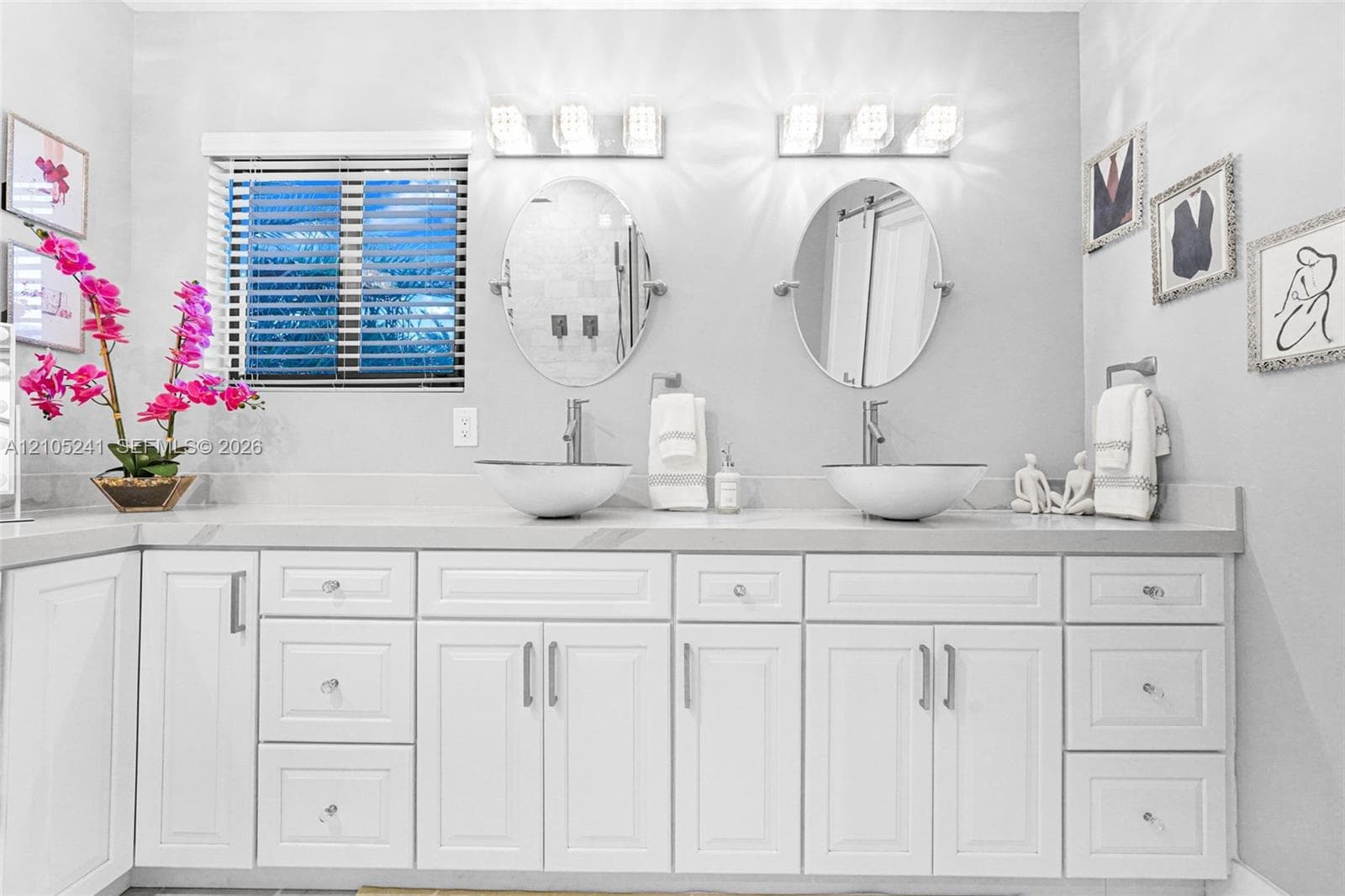 Dual vessel sinks with modern faucets, white shaker-style cabinetry, light-toned countertop, wall-mounted vanity lighting, and a horizontal window with blinds