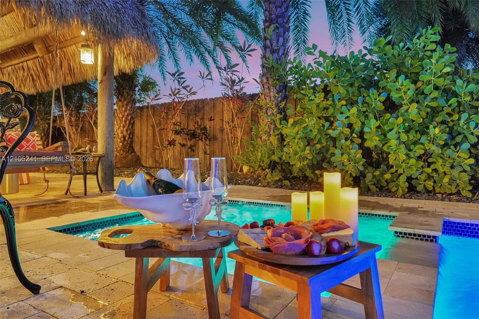 Paved patio with an adjacent swimming pool, surrounded by lush tropical landscaping and a thatched-roof structure
