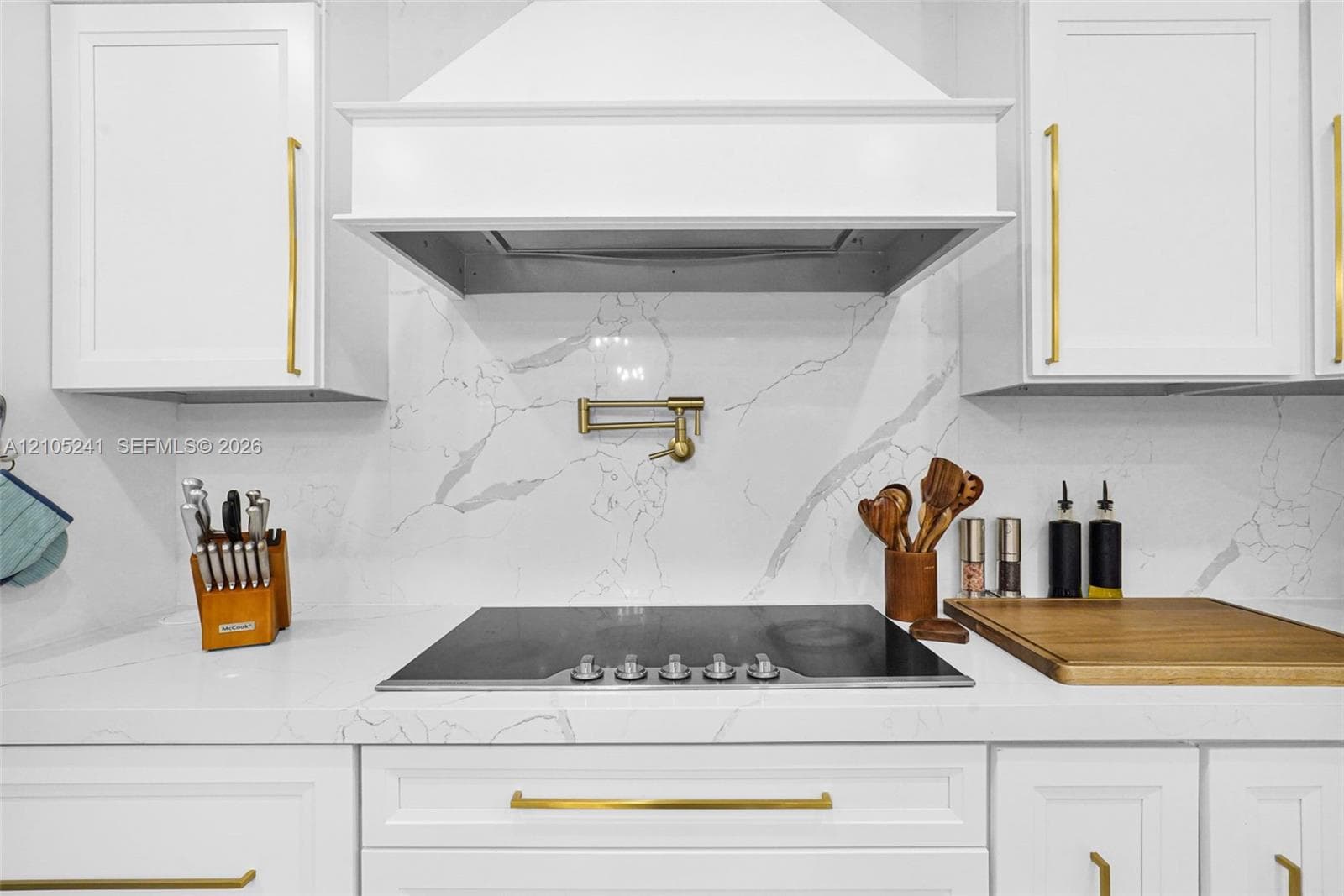 White cabinetry with gold-tone hardware, built-in cooktop, pot filler faucet, custom range hood, and stone-finish backsplash