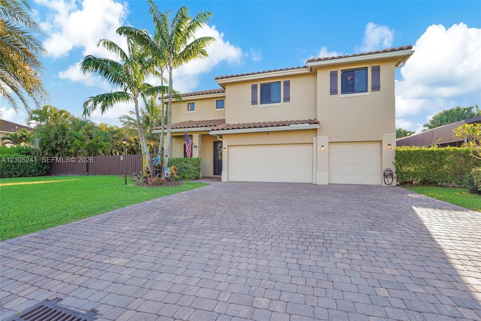 Two-story stucco exterior with tile roof, paver driveway, two garage doors, and mature palm trees