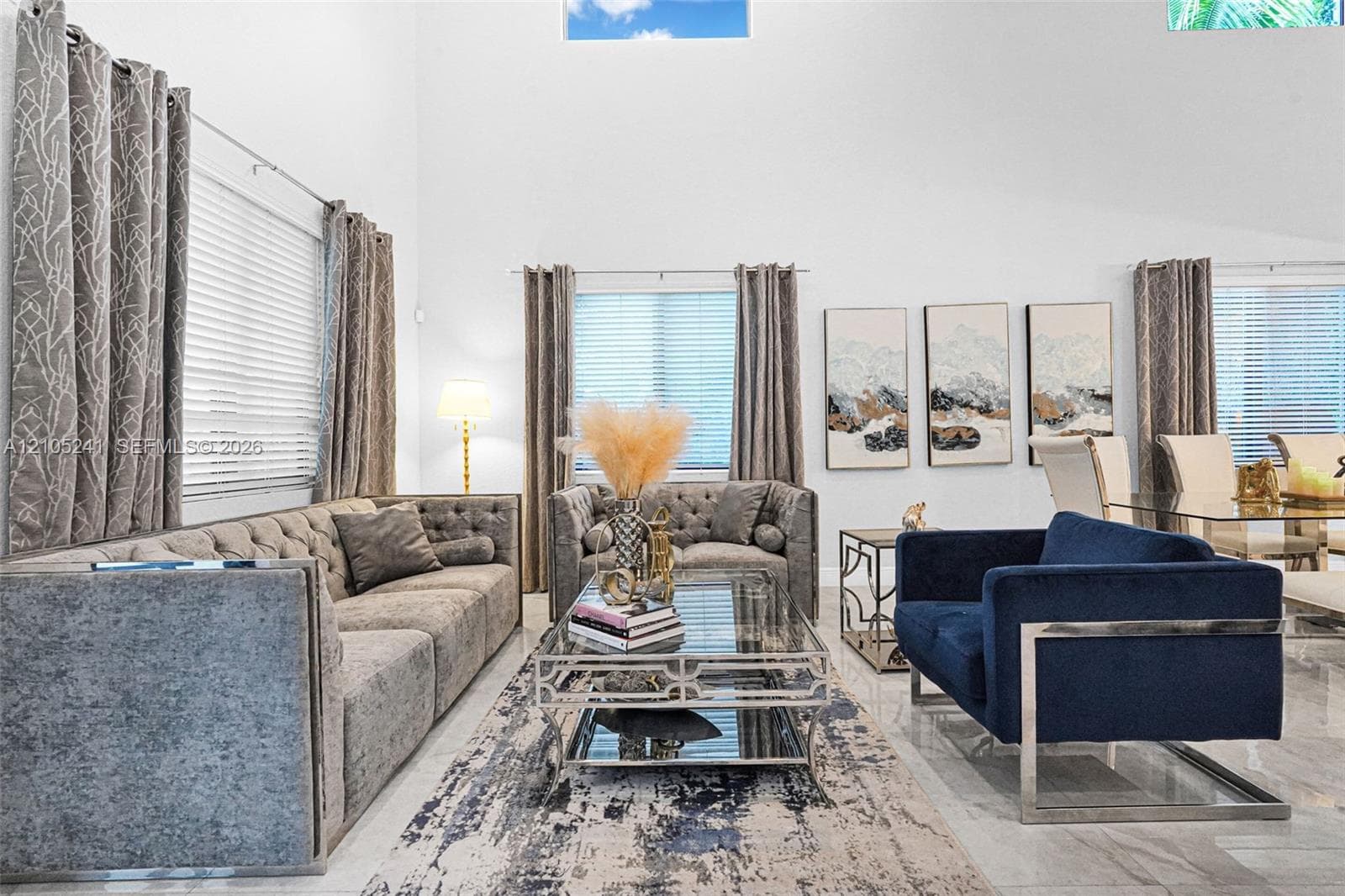 Vaulted ceiling, bright white walls, large windows with blinds, light-toned tile flooring, and a decorative area rug
