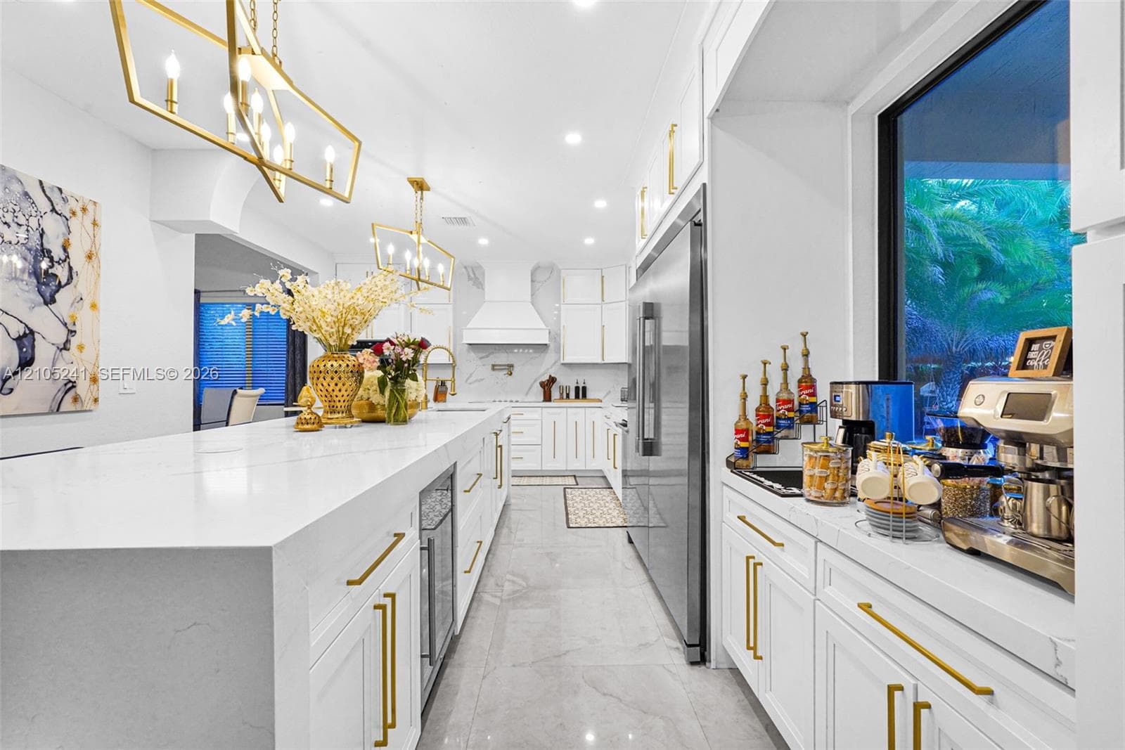 Bright kitchen featuring an extended island with a built-in beverage refrigerator, white cabinetry, stainless steel refrigerator, and gold-tone hardware