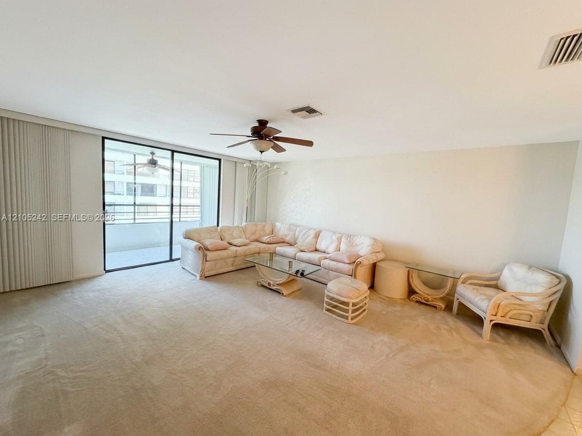 Expansive living area with neutral carpeting, vertical blinds, and a built-in ceiling fan, opening to an outdoor balcony with metal railings