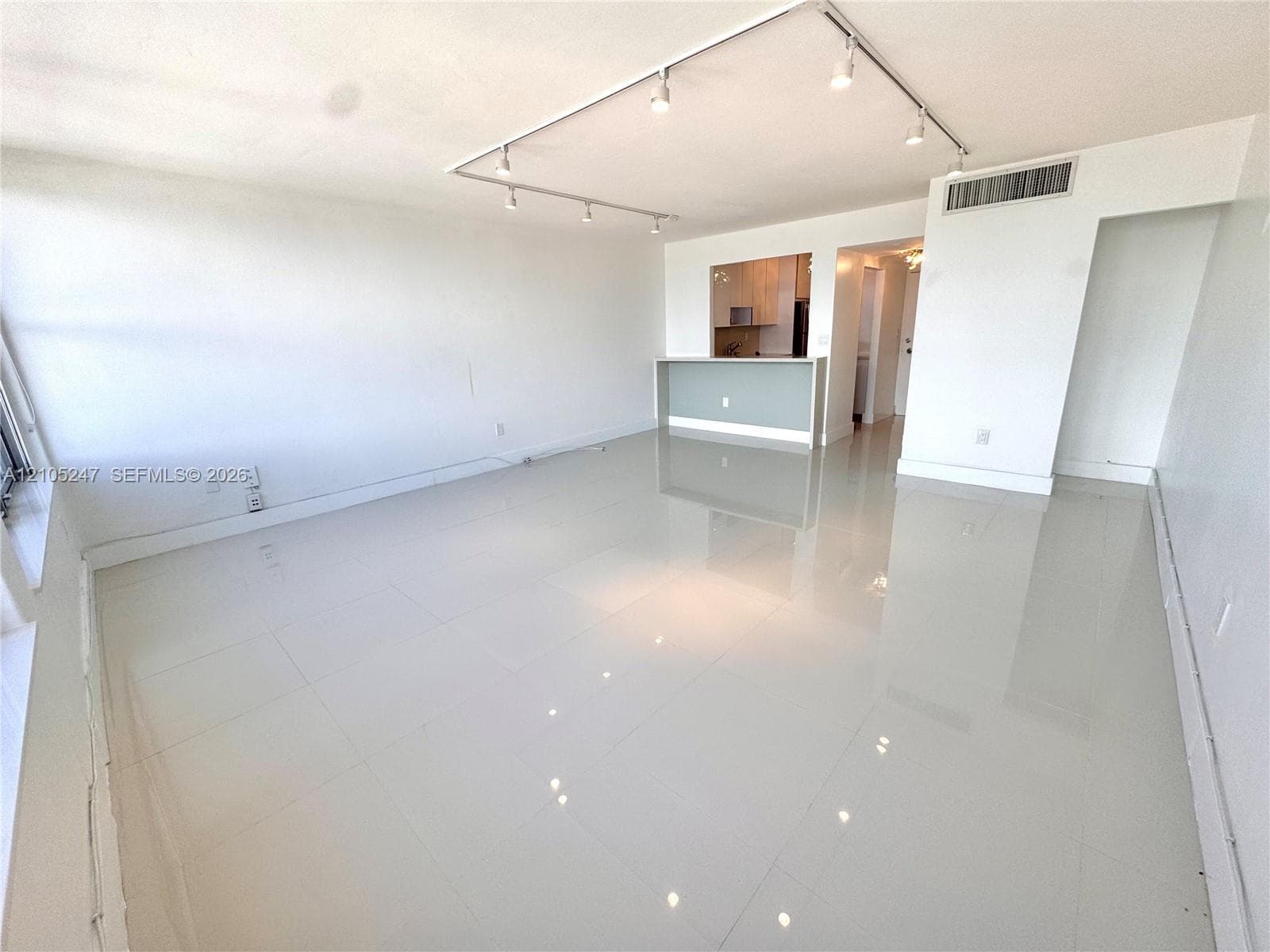 Open-concept living area featuring large format polished tile flooring, track lighting, and a partial wall separating a kitchen with light wood-finish cabinetry