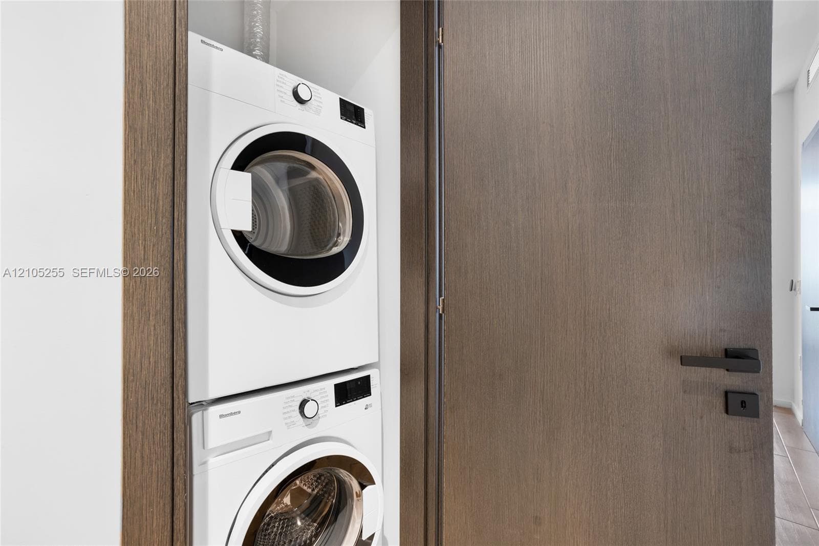 Stacked laundry center featuring high-efficiency front-load appliances housed within a recessed closet with solid wood-finish door and matte black hardware