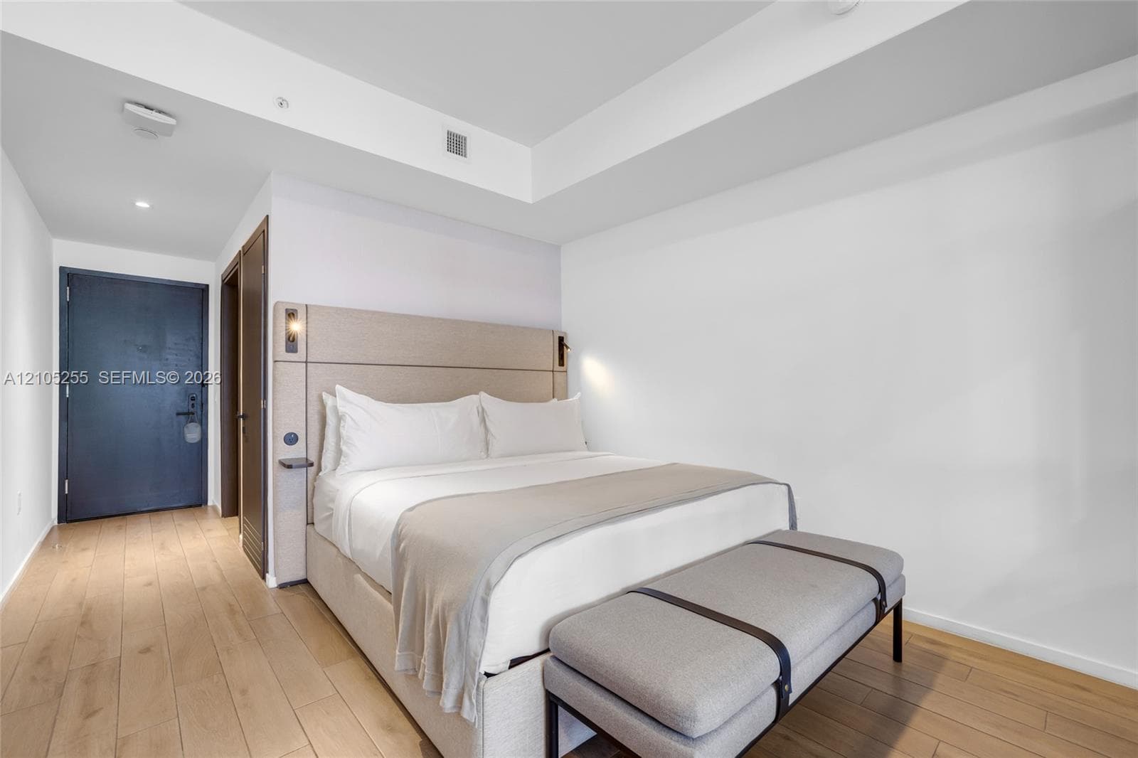 Neutral color palette featuring wood-finish flooring, tray ceiling, and a built-in upholstered headboard with integrated lighting fixtures