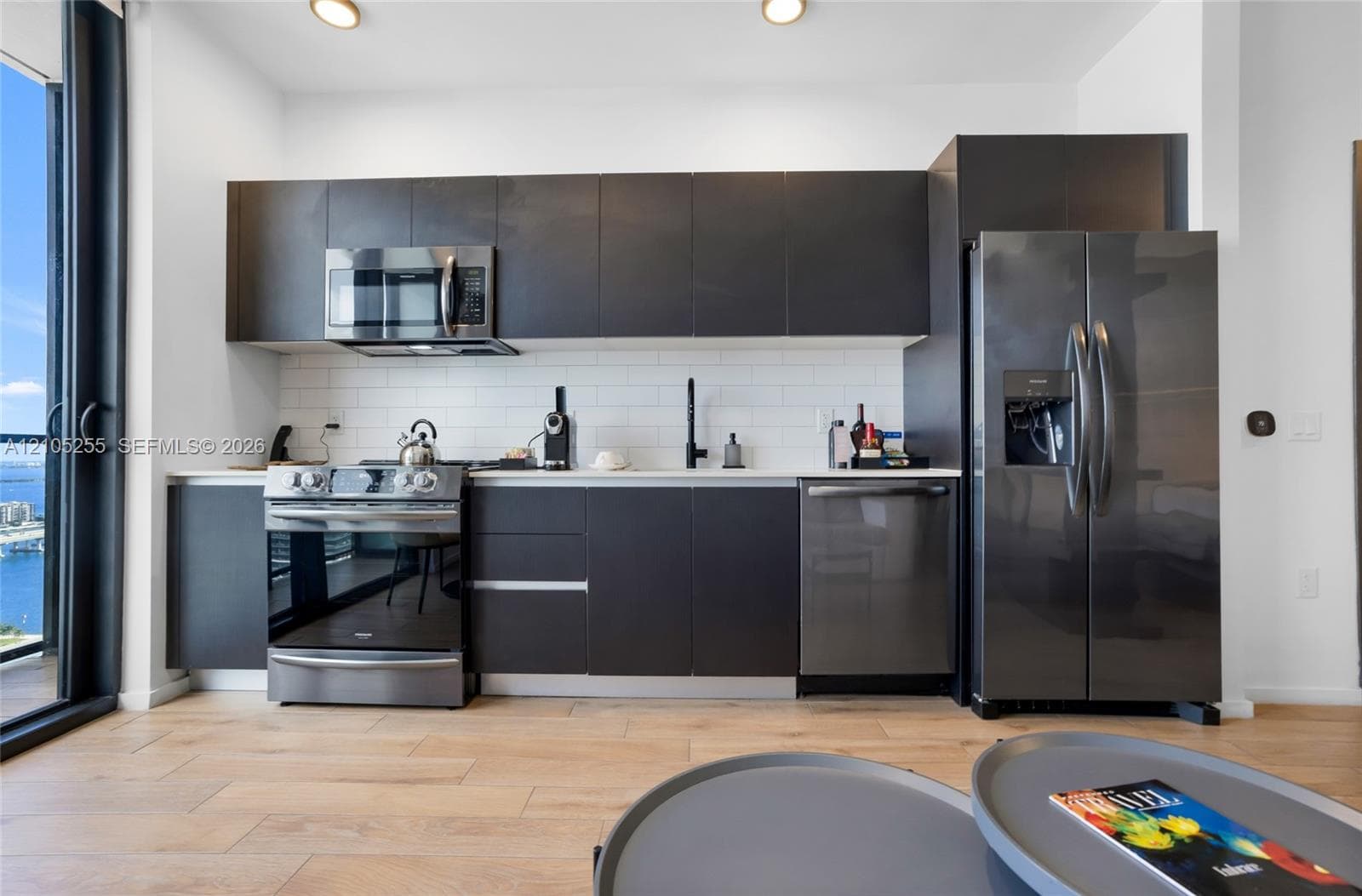 Kitchen featuring flat-panel dark cabinetry, stainless steel appliances, light-toned wood-finish flooring, and white subway tile backsplash