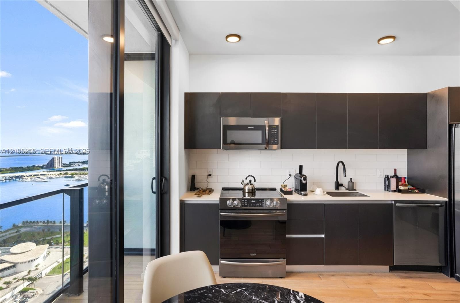 Kitchen featuring flat-panel dark cabinetry, white subway tile backsplash, stainless steel appliances, and wood-finish flooring, accompanied by floor-to-ceiling glass doors opening to a balcony