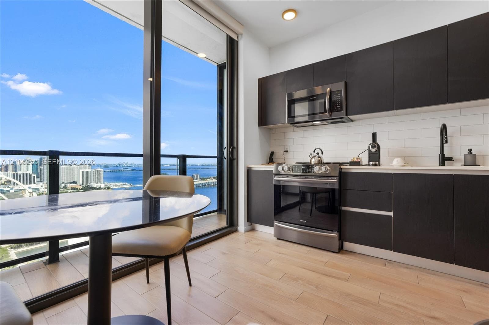 Kitchen featuring matte black flat-panel cabinetry, white subway tile backsplash, stainless steel appliances, light-toned wood-finish flooring, and floor-to-ceiling glass sliding doors