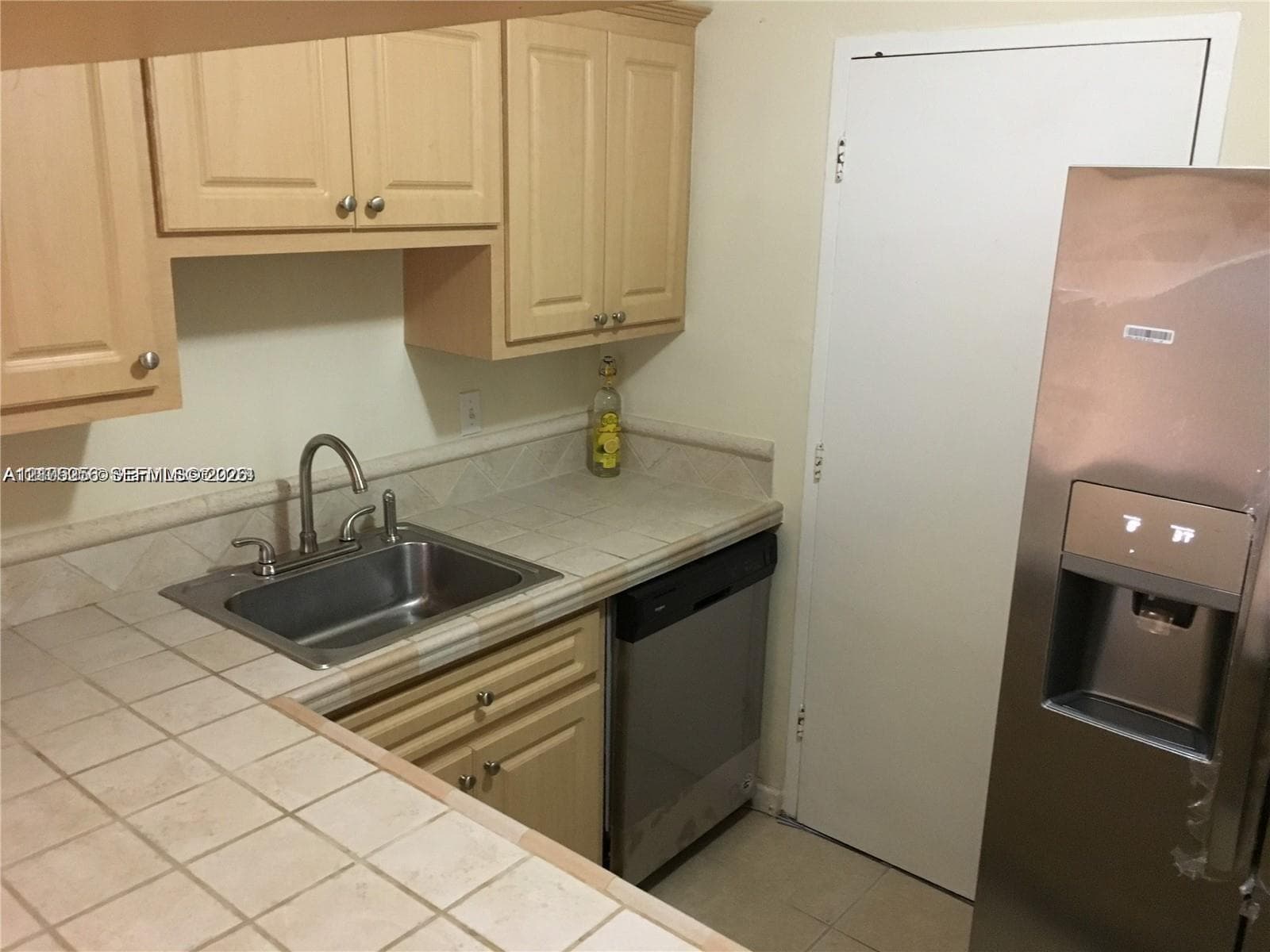 Kitchen featuring light-finish cabinetry, tiled countertops with matching backsplash, stainless steel sink with arch faucet, and a stainless steel refrigerator with external dispenser
