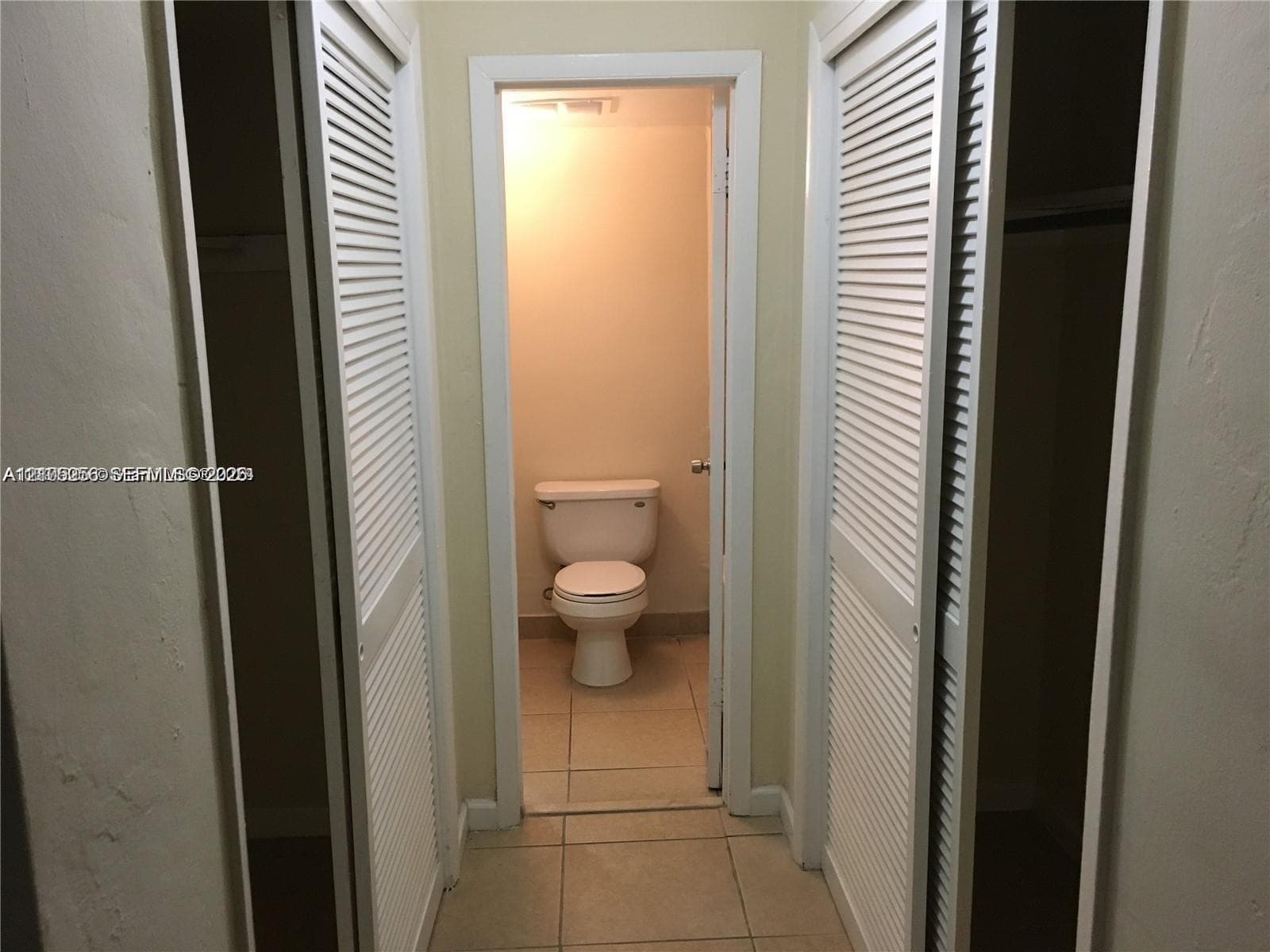 Dual closets featuring white louvered doors and large-format tile flooring leading to a water closet