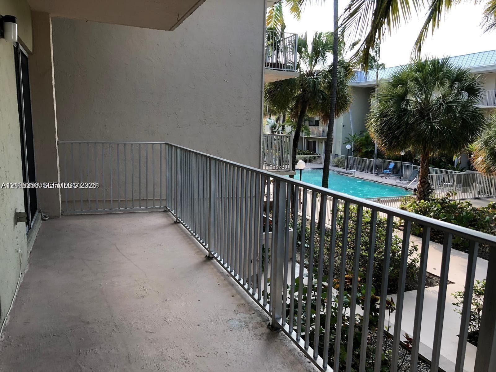 Covered balcony featuring concrete flooring and a metal railing, overlooking a swimming pool, mature palm trees, and landscaped grounds