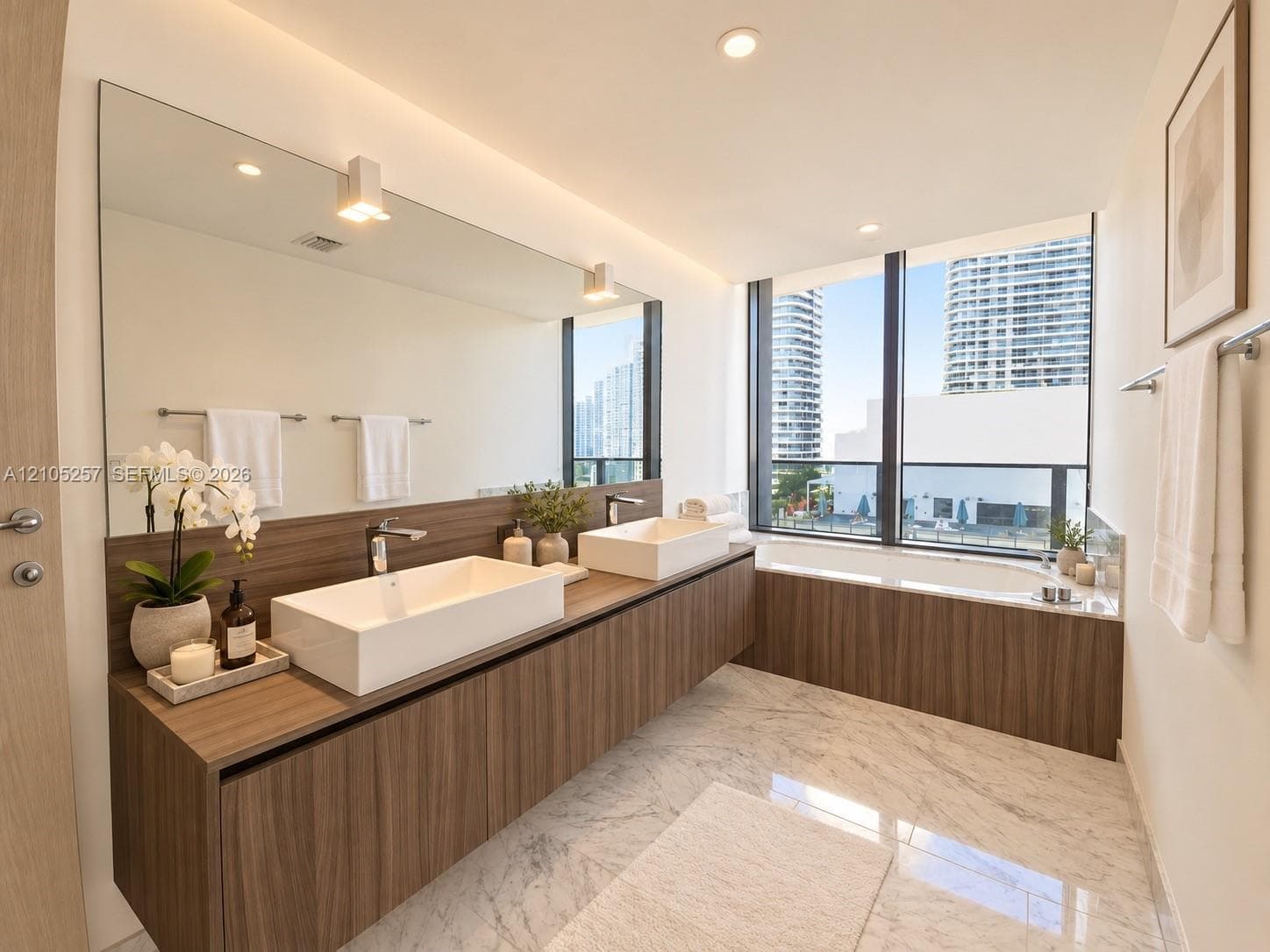 Floating wood-finish vanity with dual rectangular vessel sinks, floor-to-ceiling windows, built-in soaking tub with wood-finish surround, and stone-finish tile flooring