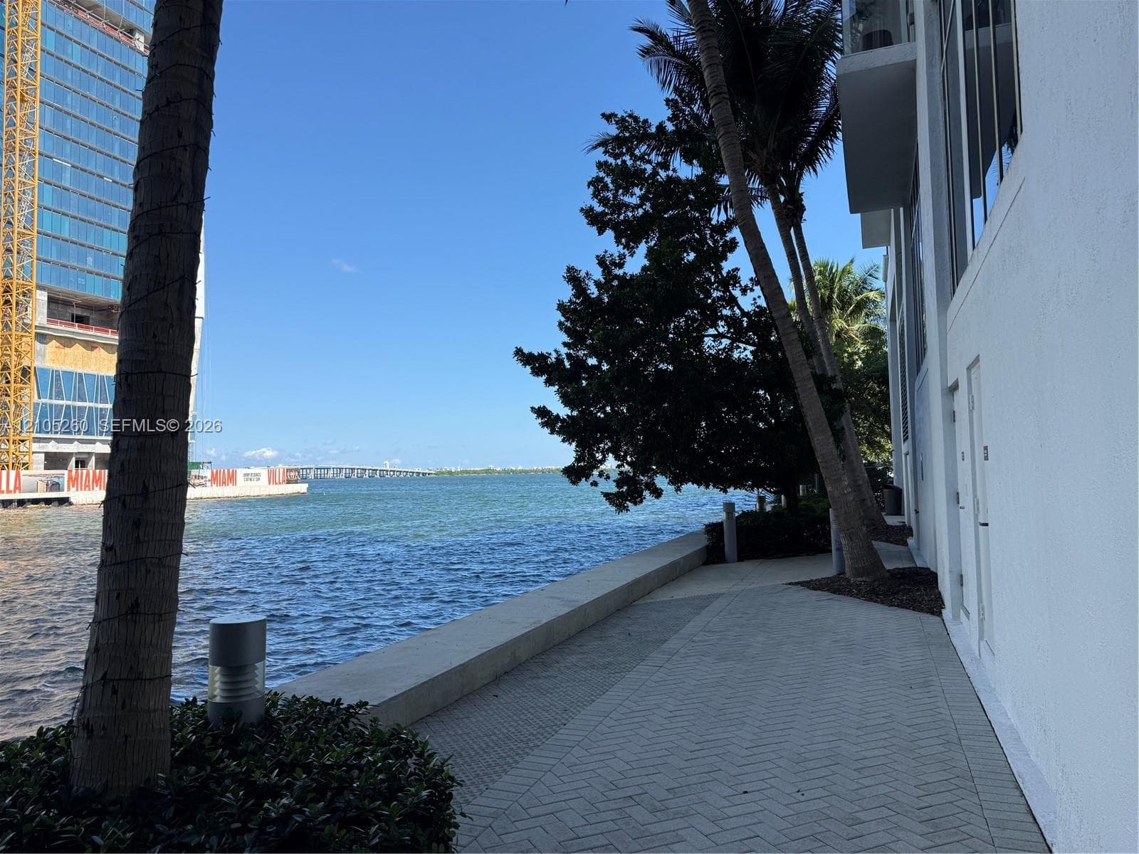 Waterfront paved walkway featuring herringbone pattern masonry, concrete retaining wall, mature palm trees, and structural landscape lighting