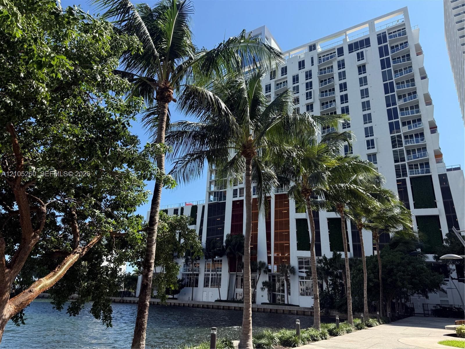 High-rise residential building featuring a white facade, staggered private balconies, multi-story vertical paneled accent walls, and waterfront positioning with mature palm landscaping