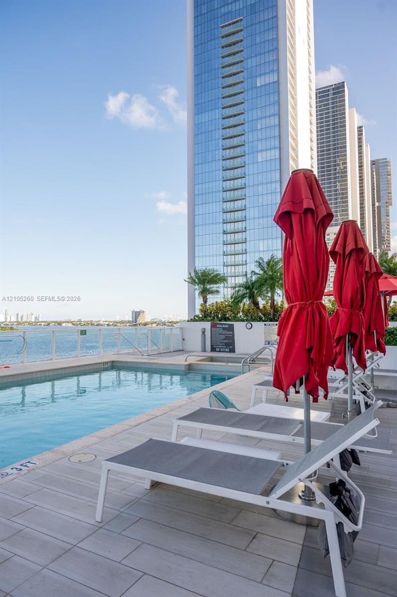 Outdoor pool area with a water body backdrop, multiple lounge chairs, and a contemporary high-rise building