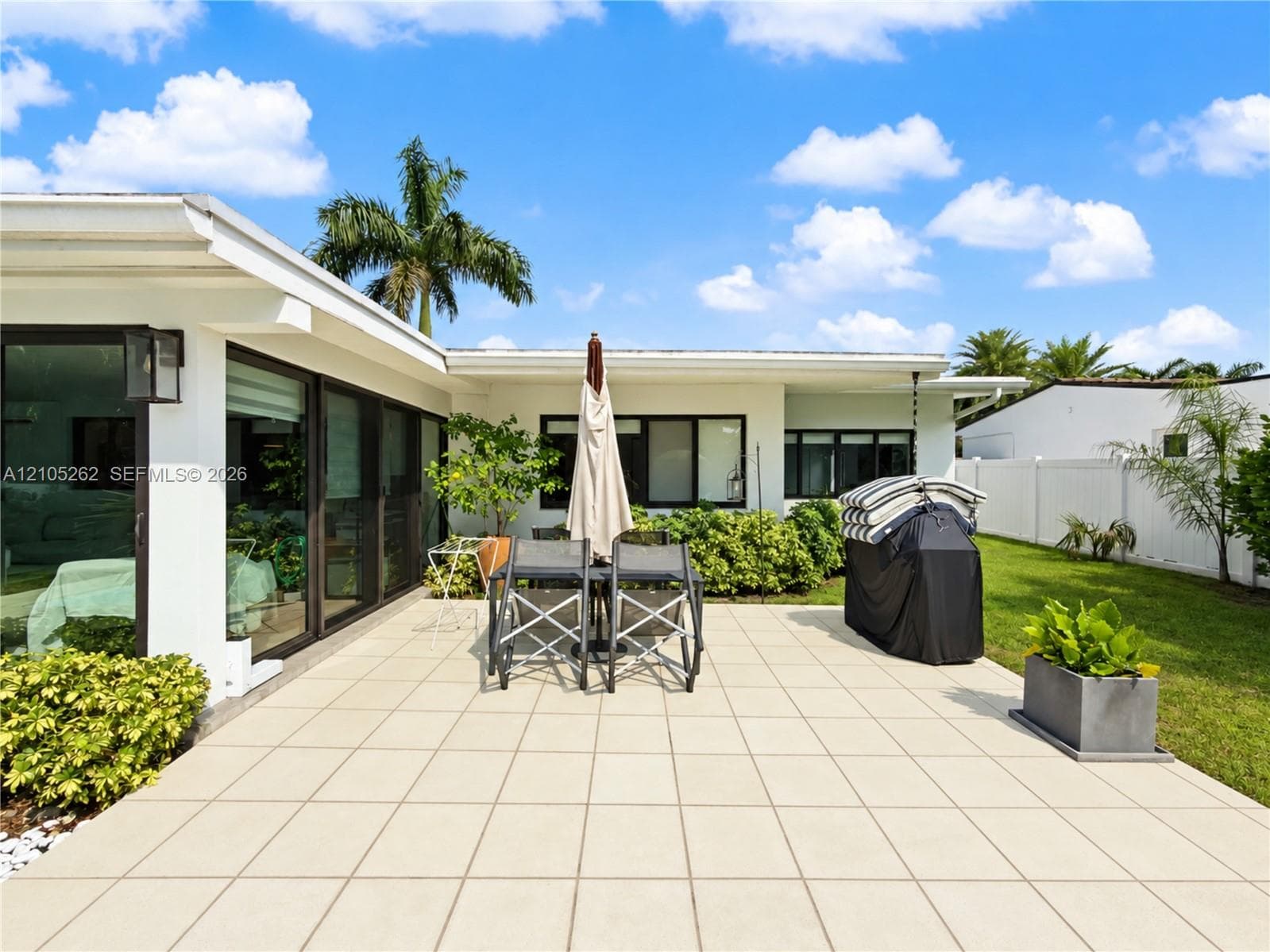 Expansive patio with large format square tiles, surrounded by mature landscaping, and featuring a white perimeter fence