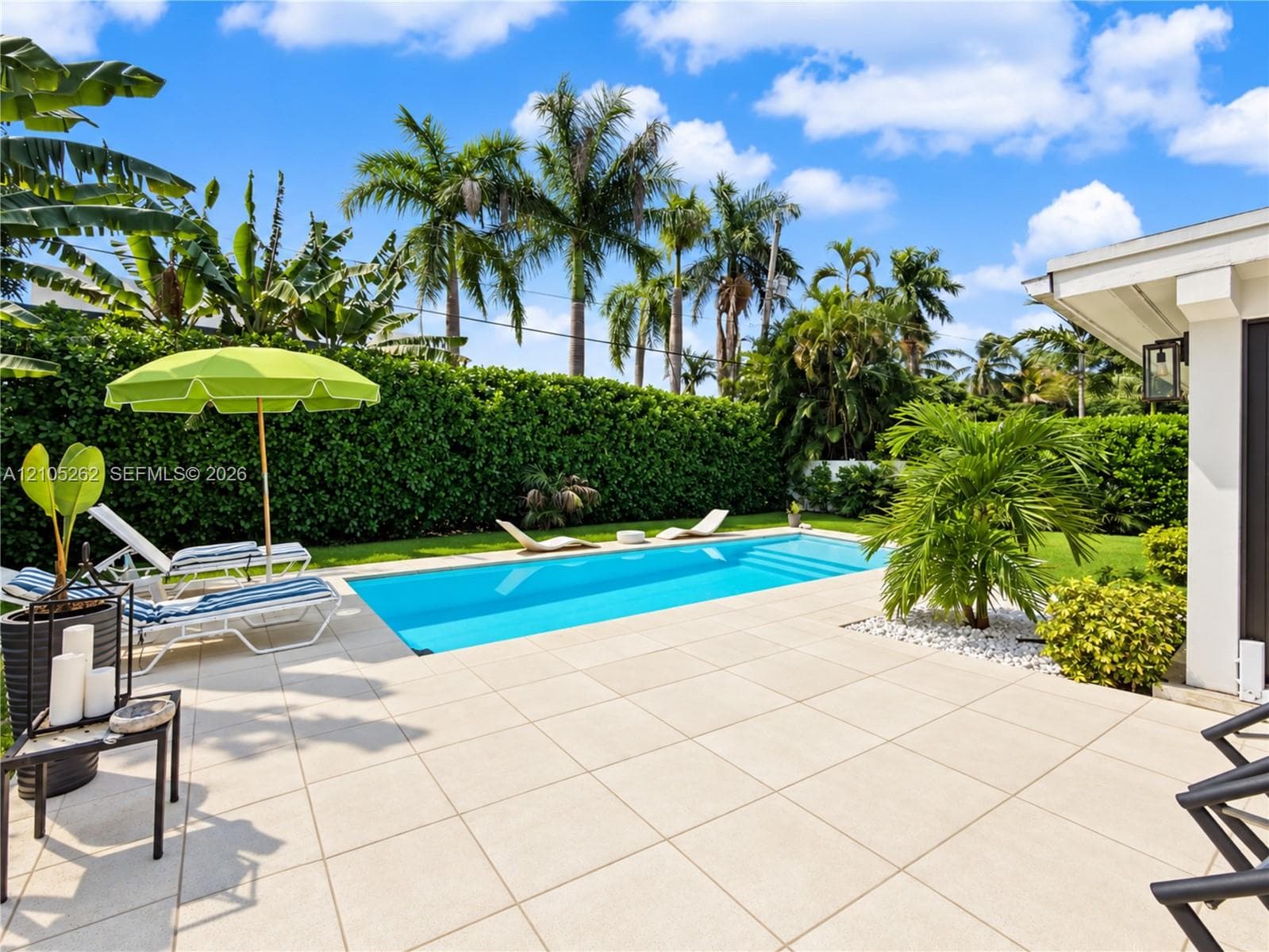Outdoor swimming pool with a large tiled patio surround, bordered by manicured hedges and mature palm trees