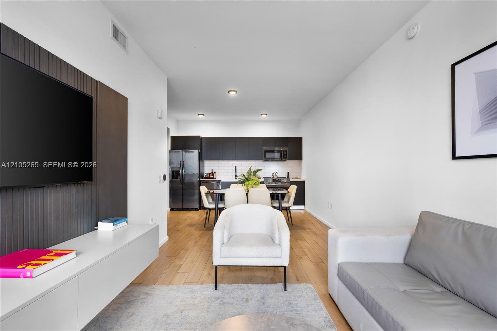 Modern interior featuring wood-finish flooring, a matte-finish accent wall with a wall-mounted media console, dark cabinetry, and white tile backsplash