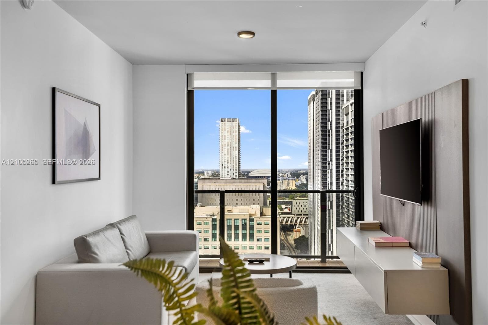 Floor-to-ceiling windows, integrated wall-mounted media console with wood-finish paneling, and private balcony with urban skyline scenery