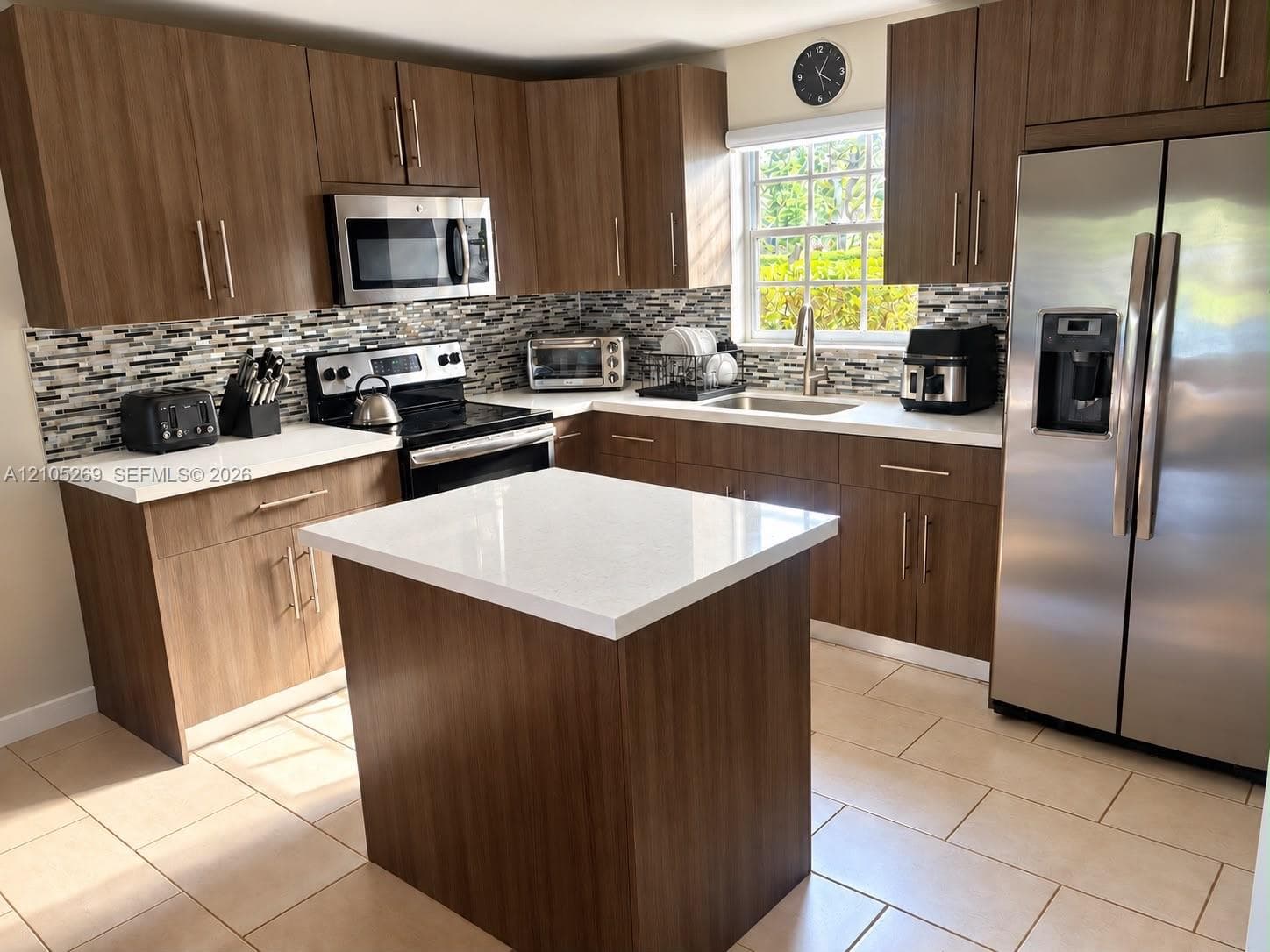 Kitchen featuring wood-finish cabinetry, stainless steel appliances, a center island with a white countertop, a mosaic tile backsplash, and light-toned tile flooring