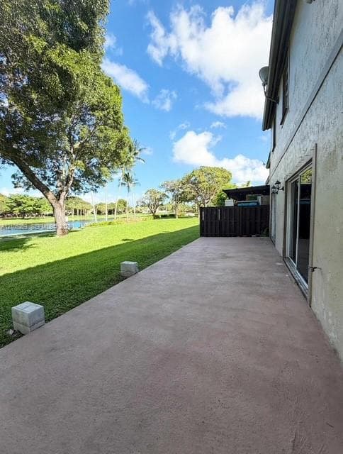 Expansive concrete patio with sliding glass doors, a manicured lawn, mature trees, and a water feature