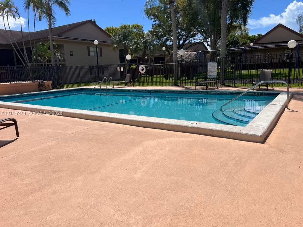 Community pool with a surrounding black metal fence, ample deck space, and mature palm trees