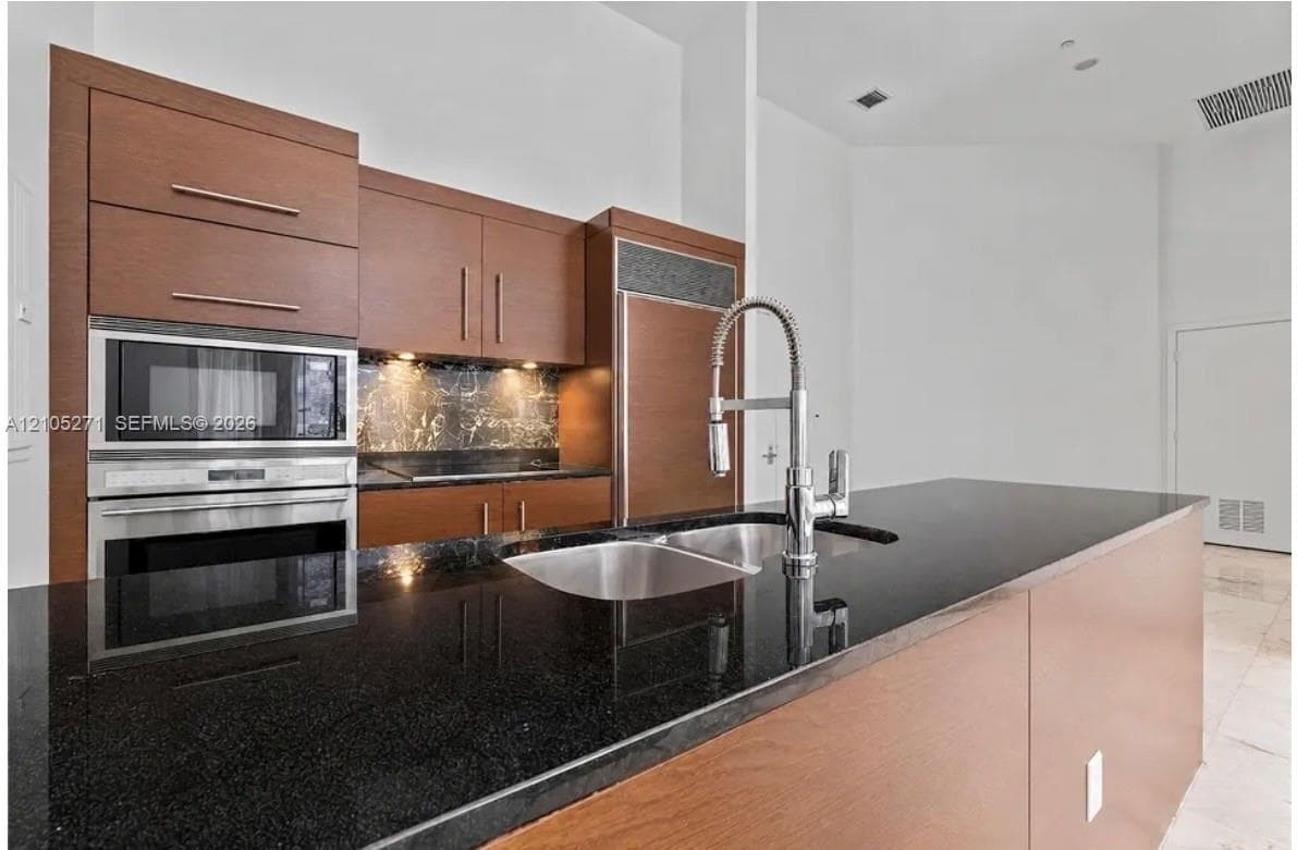Kitchen featuring flat-panel wood cabinetry, black stone countertops, built-in stainless steel wall oven and microwave, panel-ready refrigerator, and a stainless steel gooseneck faucet