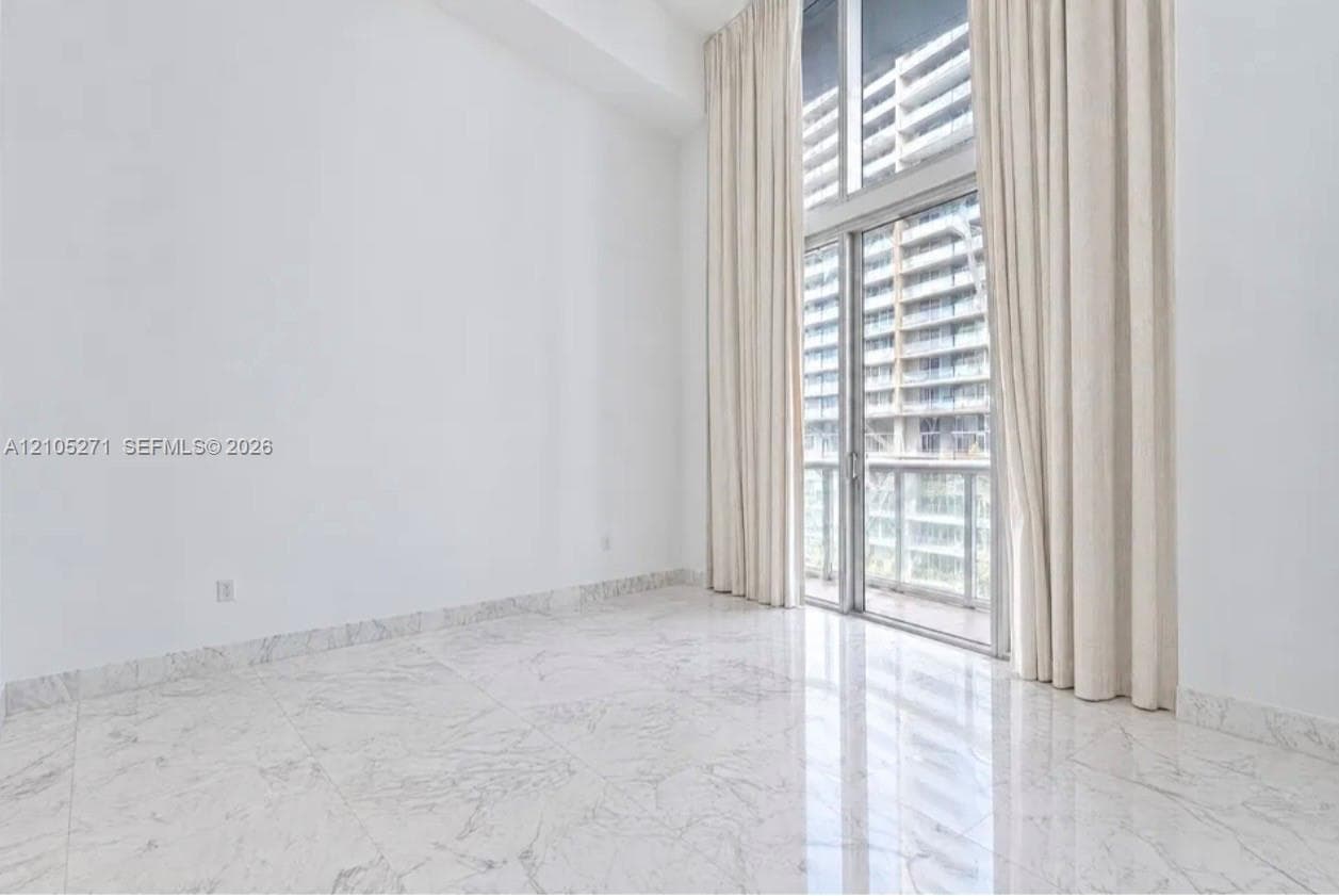 High-ceilinged room featuring polished stone-finish floor tiling, floor-to-ceiling glass paneling, and access to a balcony with glass railing