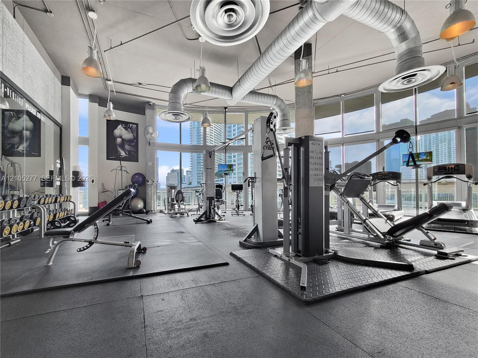 Fitness center with full-height windows, exposed ductwork, rubber flooring, and industrial pendant lighting