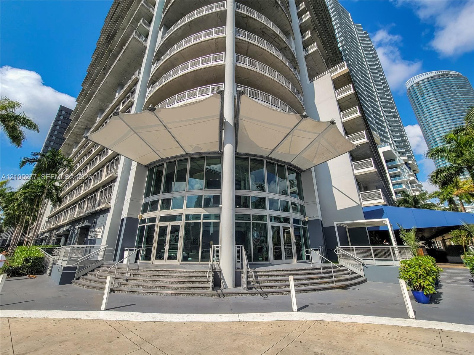 Modern high-rise building featuring a curved glass facade, multiple balconies, a fabric canopy entrance, and mature palm trees