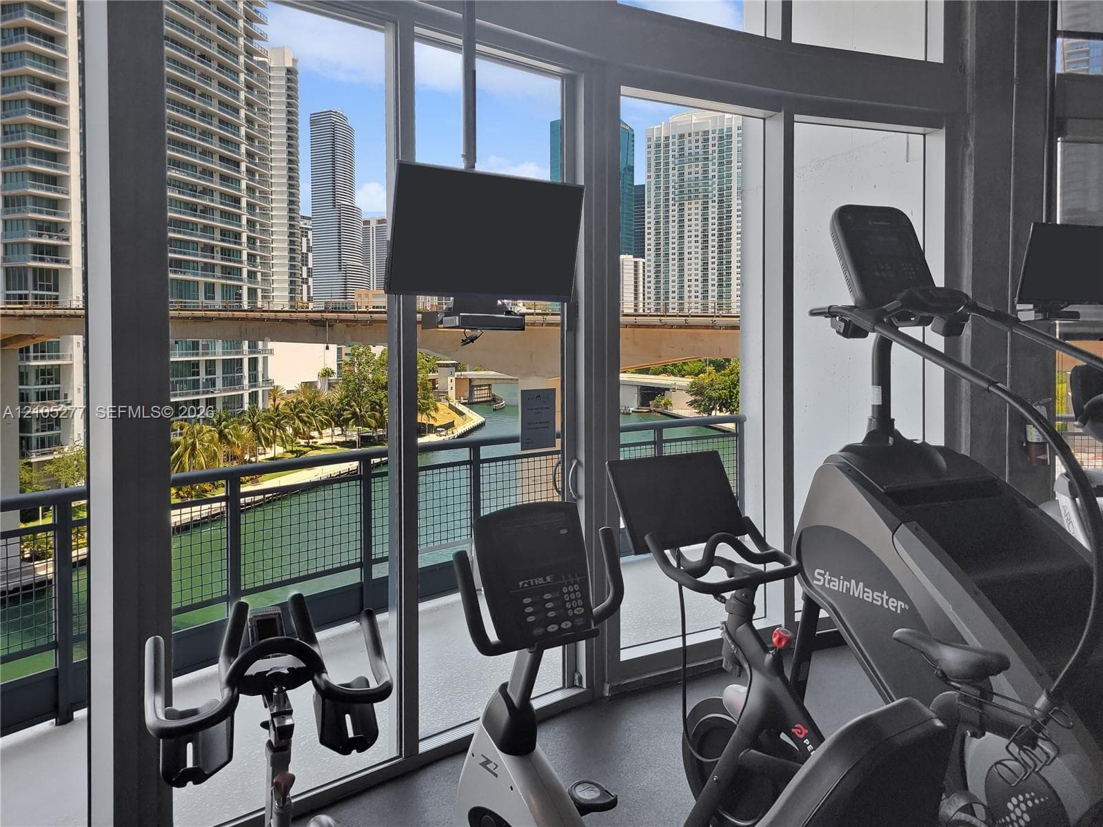 Fitness area with floor-to-ceiling windows, multiple exercise machines, and a balcony with metal railing overlooking a waterway