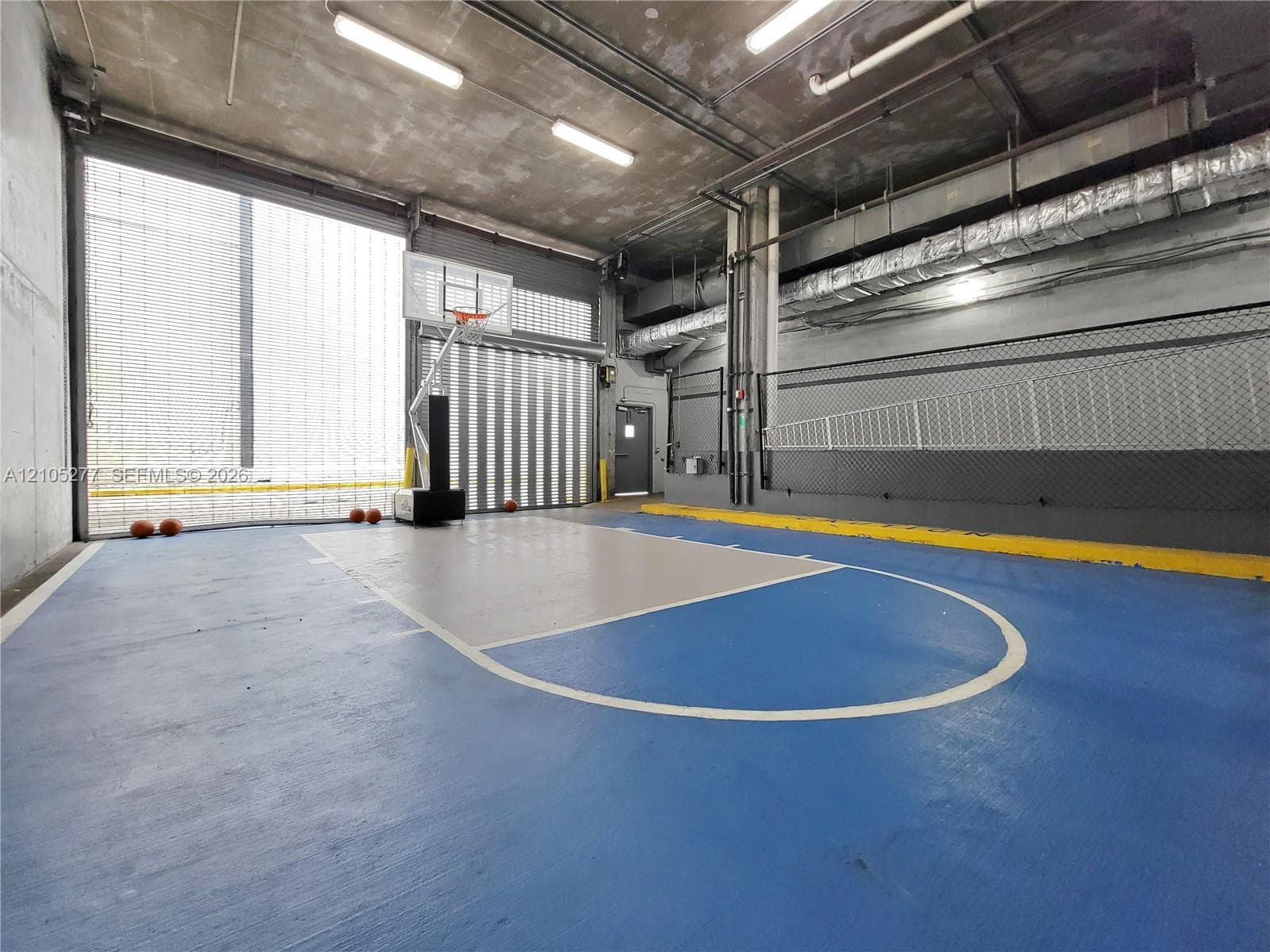 Indoor basketball court featuring blue and gray flooring with white court lines, concrete walls, and overhead lighting