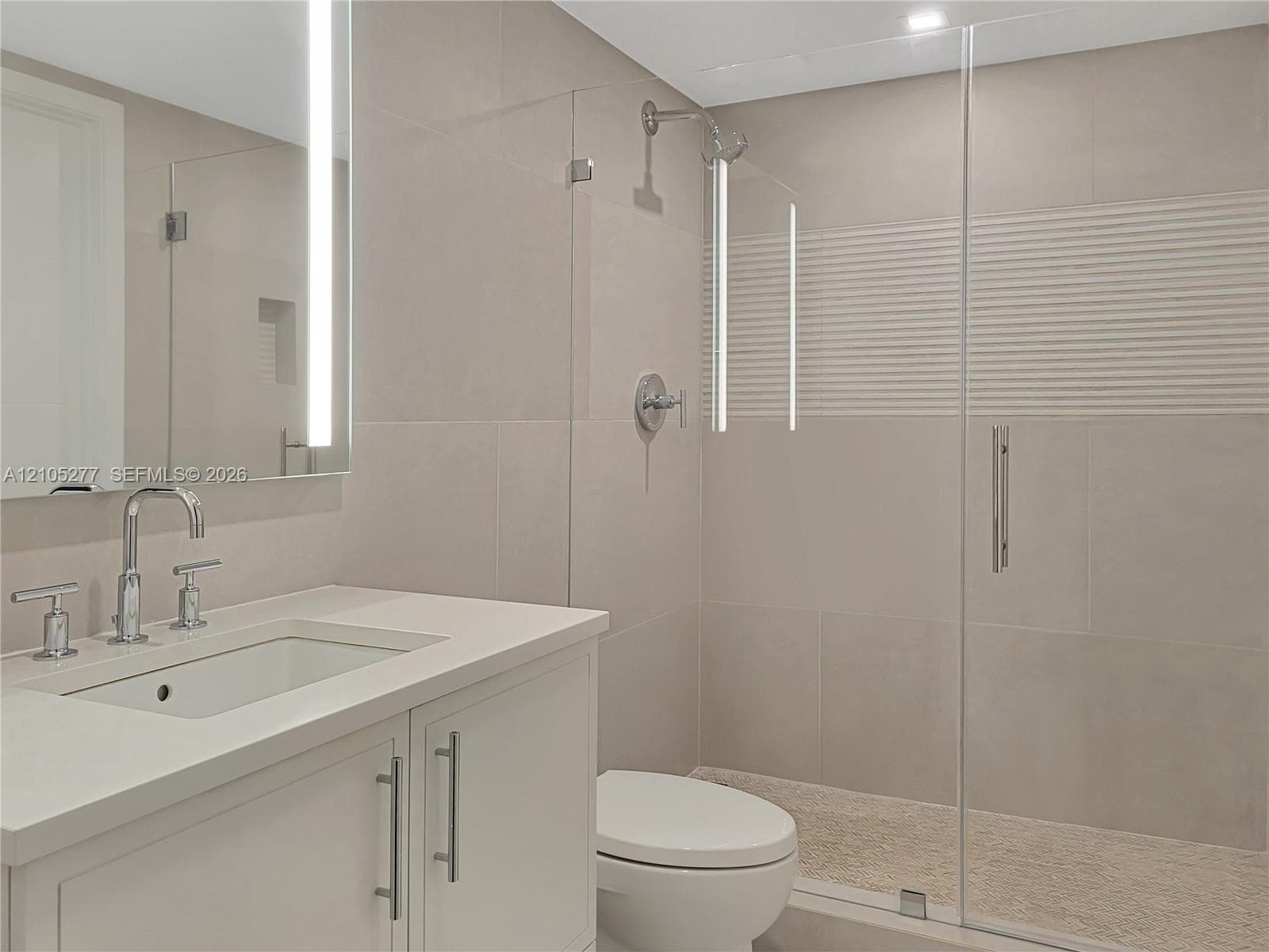Vanity with integrated sink and chrome faucet, back-lit mirror, glass-enclosed shower with rain head, textured accent tile, and flush-mounted ceiling lighting